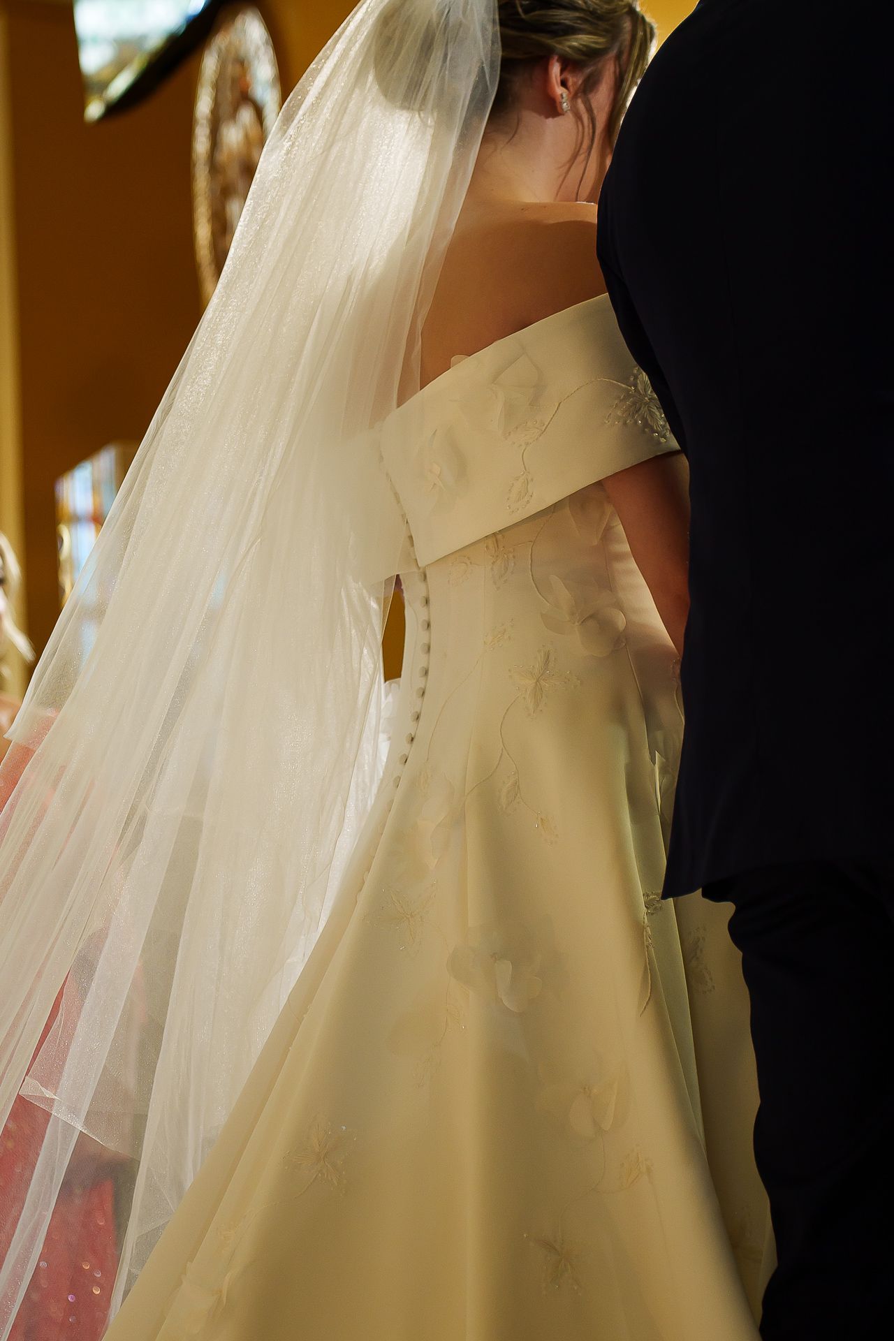 Bride in elegant off-shoulder white wedding gown with delicate veil, viewed from behind during ceremony.
