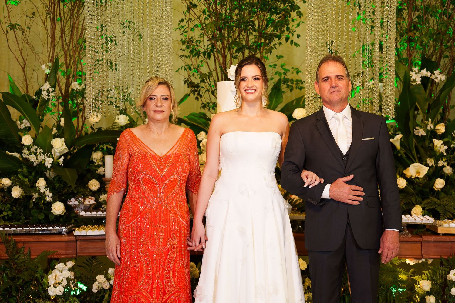 Bride in white gown poses with parents in elegant wedding venue adorned with white flowers and greenery.