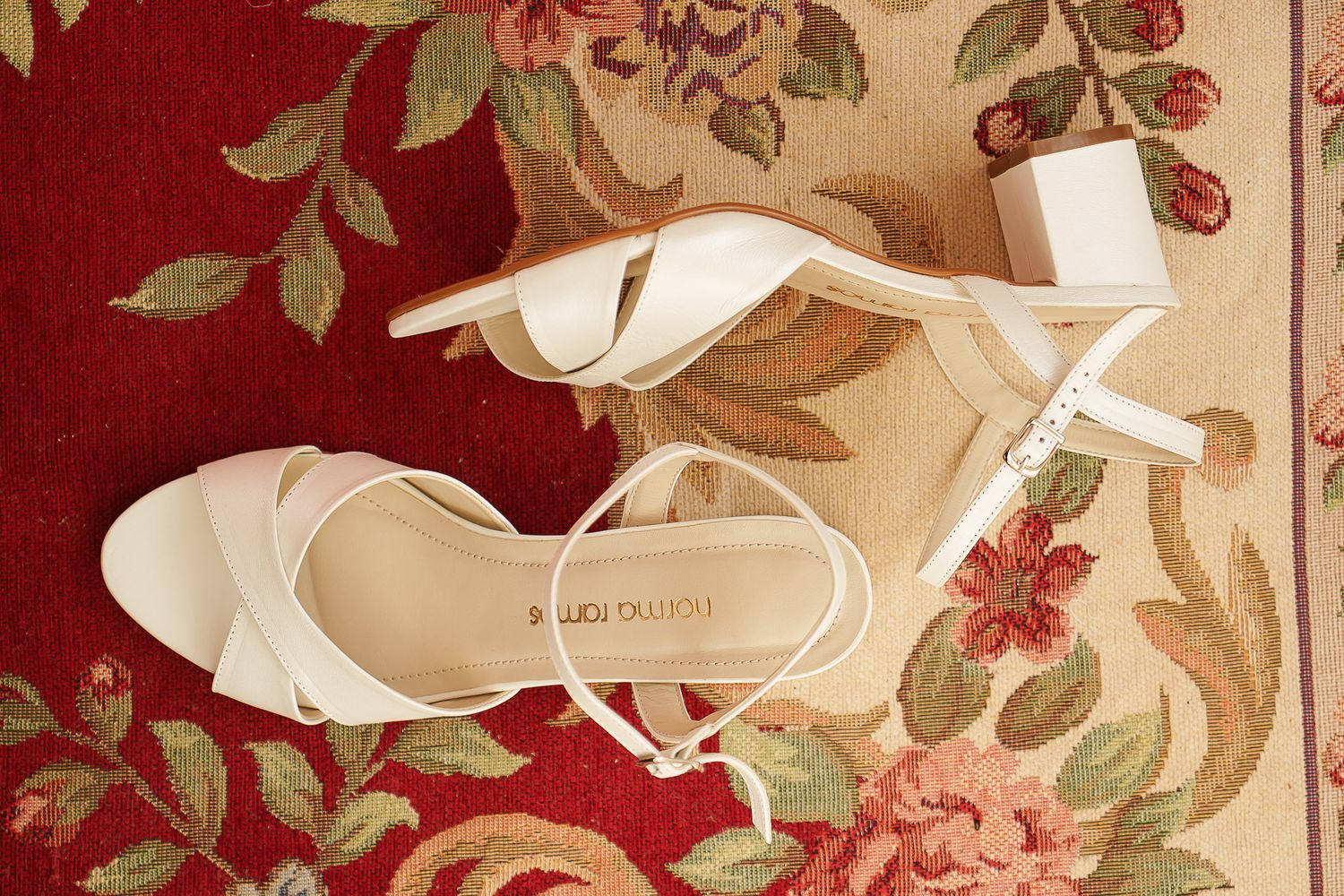 White bridal heeled sandals by Marco Oliva displayed on a floral red carpet.