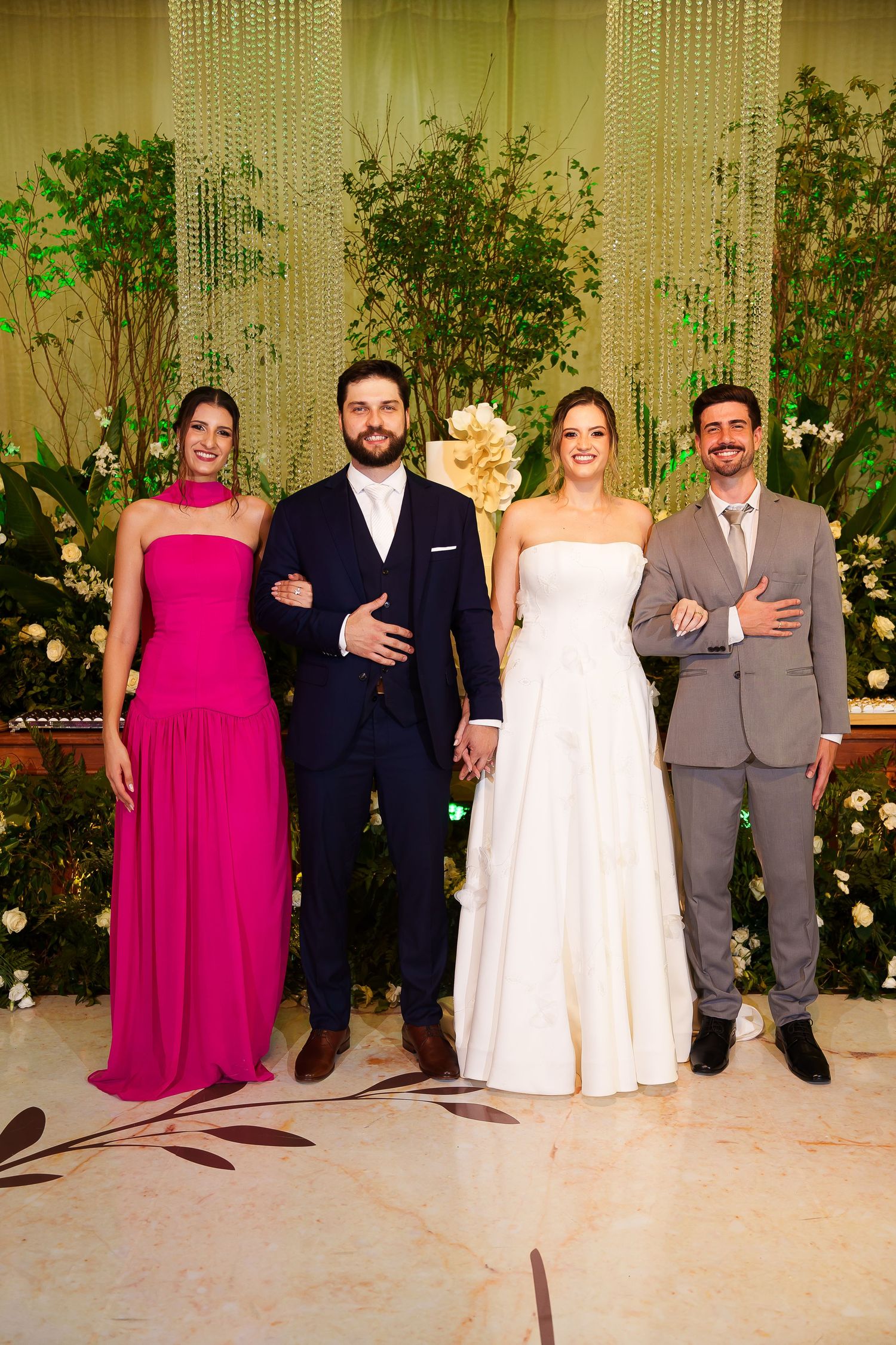Wedding party of four posing elegantly, bride in white gown, woman in pink dress, grooms in navy and gray suits.