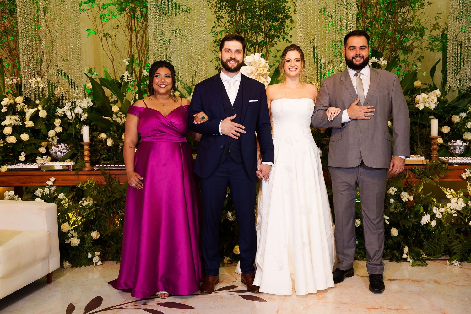 Wedding party of four posing elegantly in formal attire before a lush floral backdrop at a reception venue.