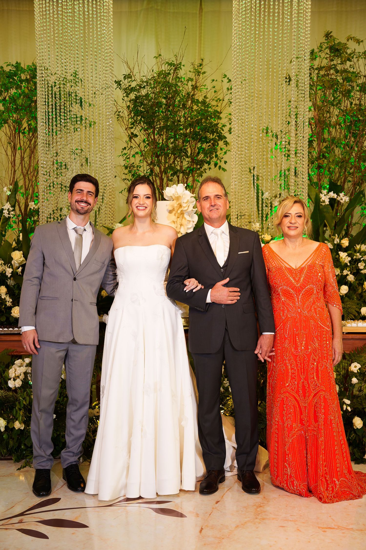 Wedding party posing elegantly, bride in white gown with groom and family in formal attire before floral backdrop.