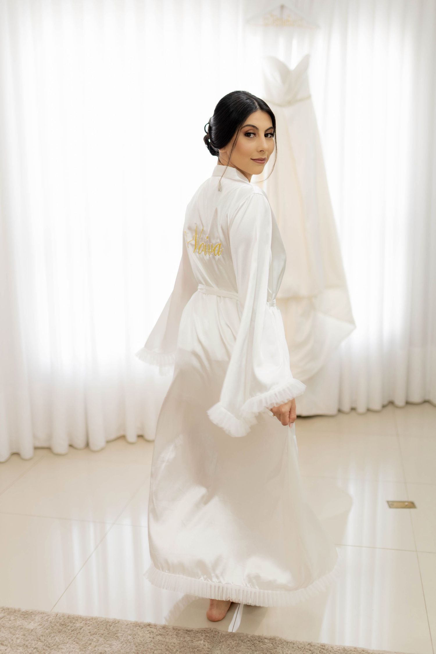 Bride in white satin robe with gold embroidery poses elegantly before her wedding gown hanging by sheer curtains.