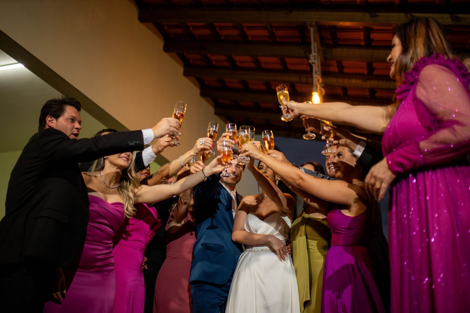 Wedding toast with bride in white, groom in blue, and bridesmaids in pink gowns raising champagne glasses together.