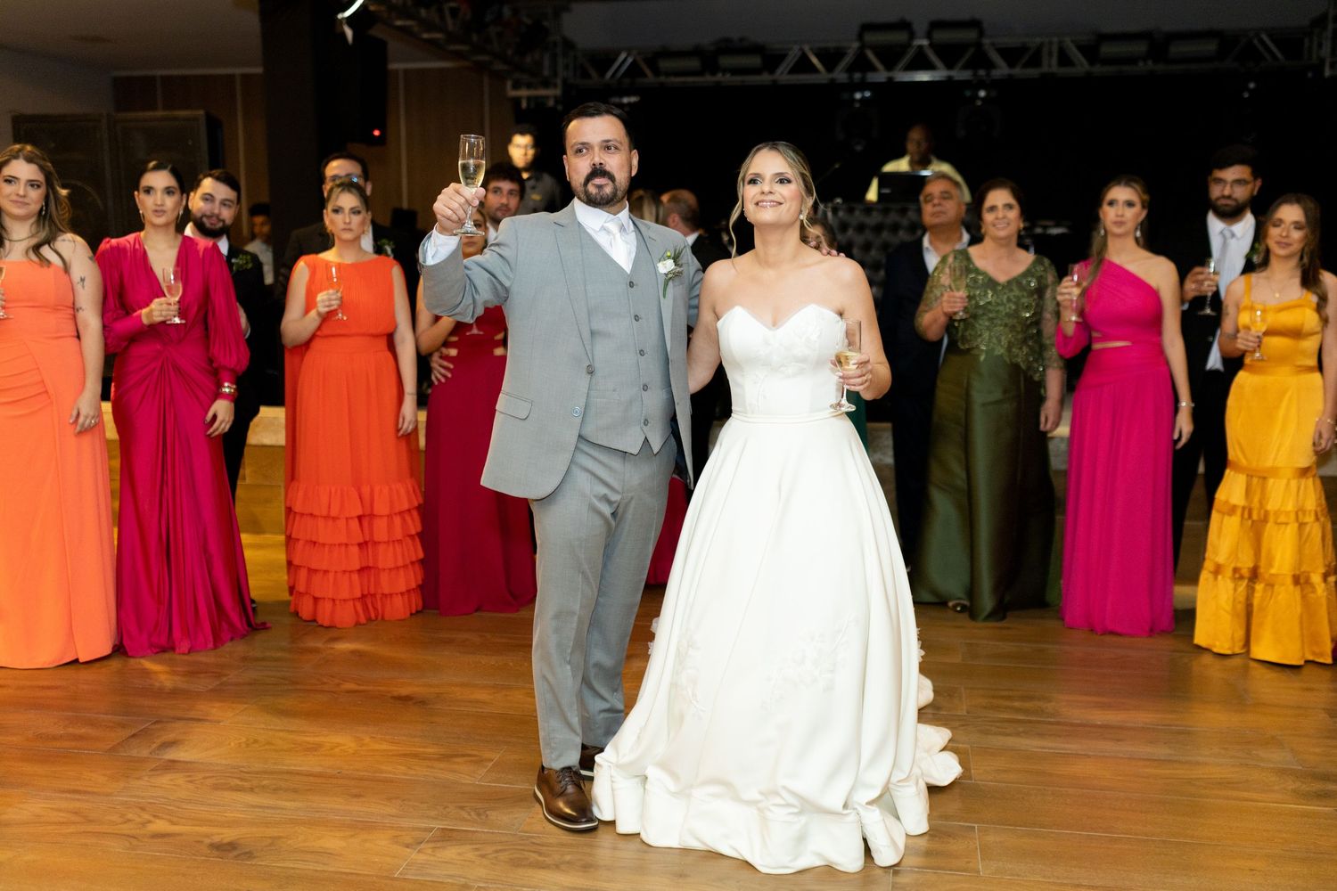 Bride and groom toast champagne on wedding dance floor surrounded by guests in colorful formal gowns.