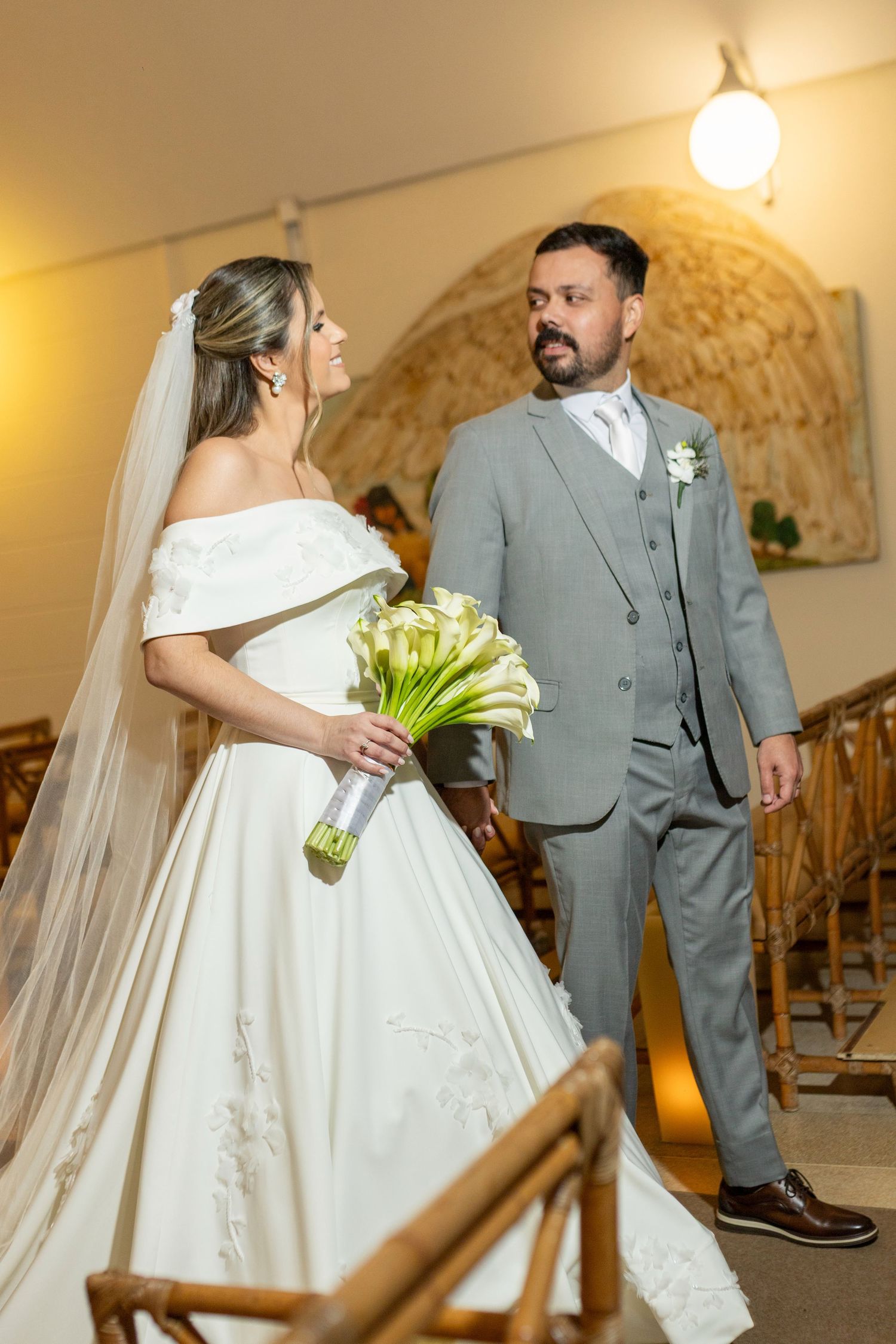 Bride in white off-shoulder gown holding calla lily bouquet stands with groom in gray suit inside elegant wedding venue.