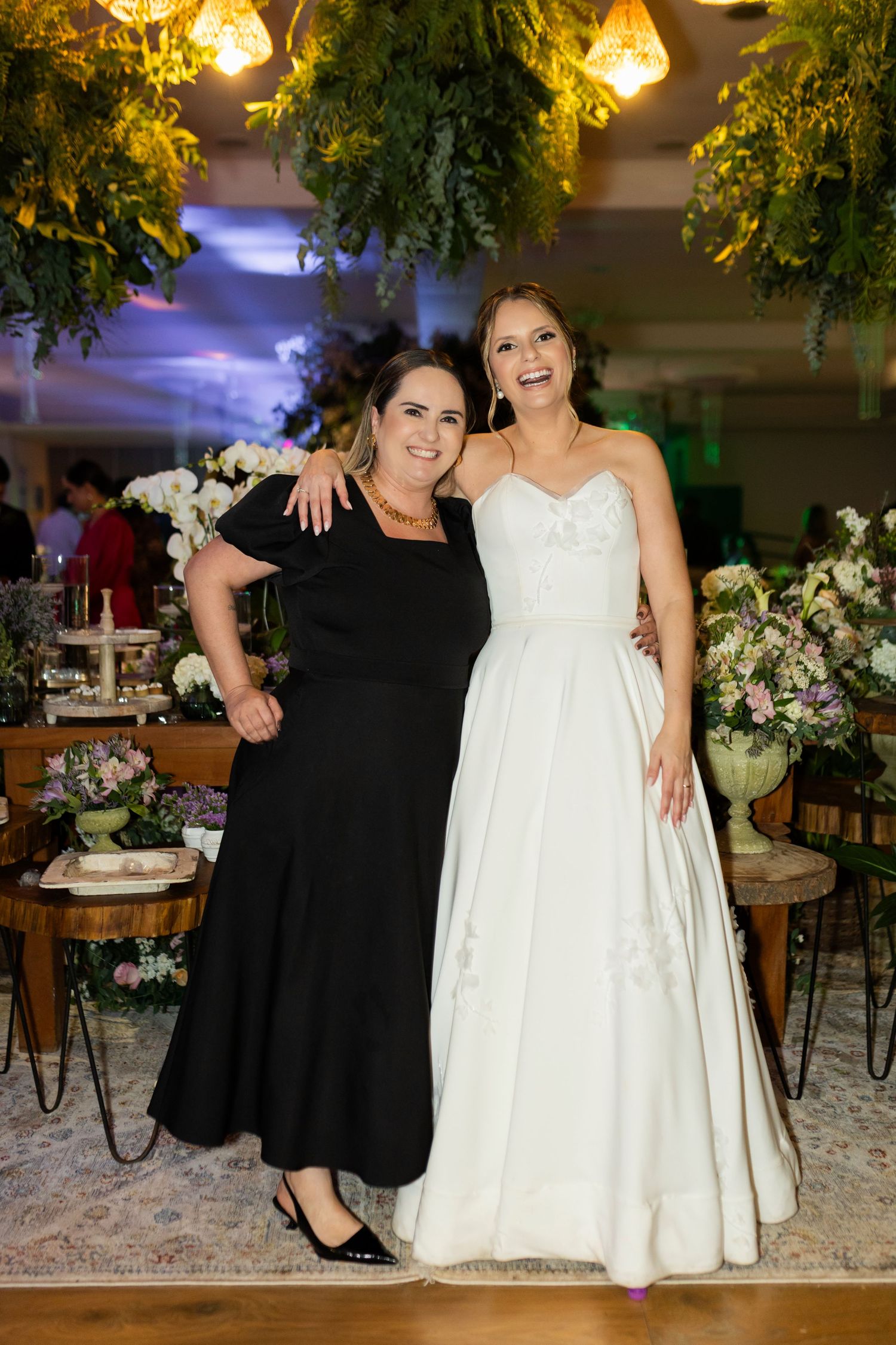 A bride in a white gown poses with a woman in a black dress at an elegantly decorated wedding reception venue.