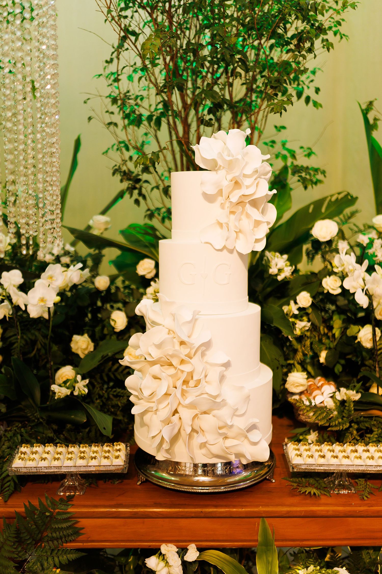 Elegant four-tier white wedding cake adorned with large sugar flowers, displayed on a wooden table surrounded by lush greenery.