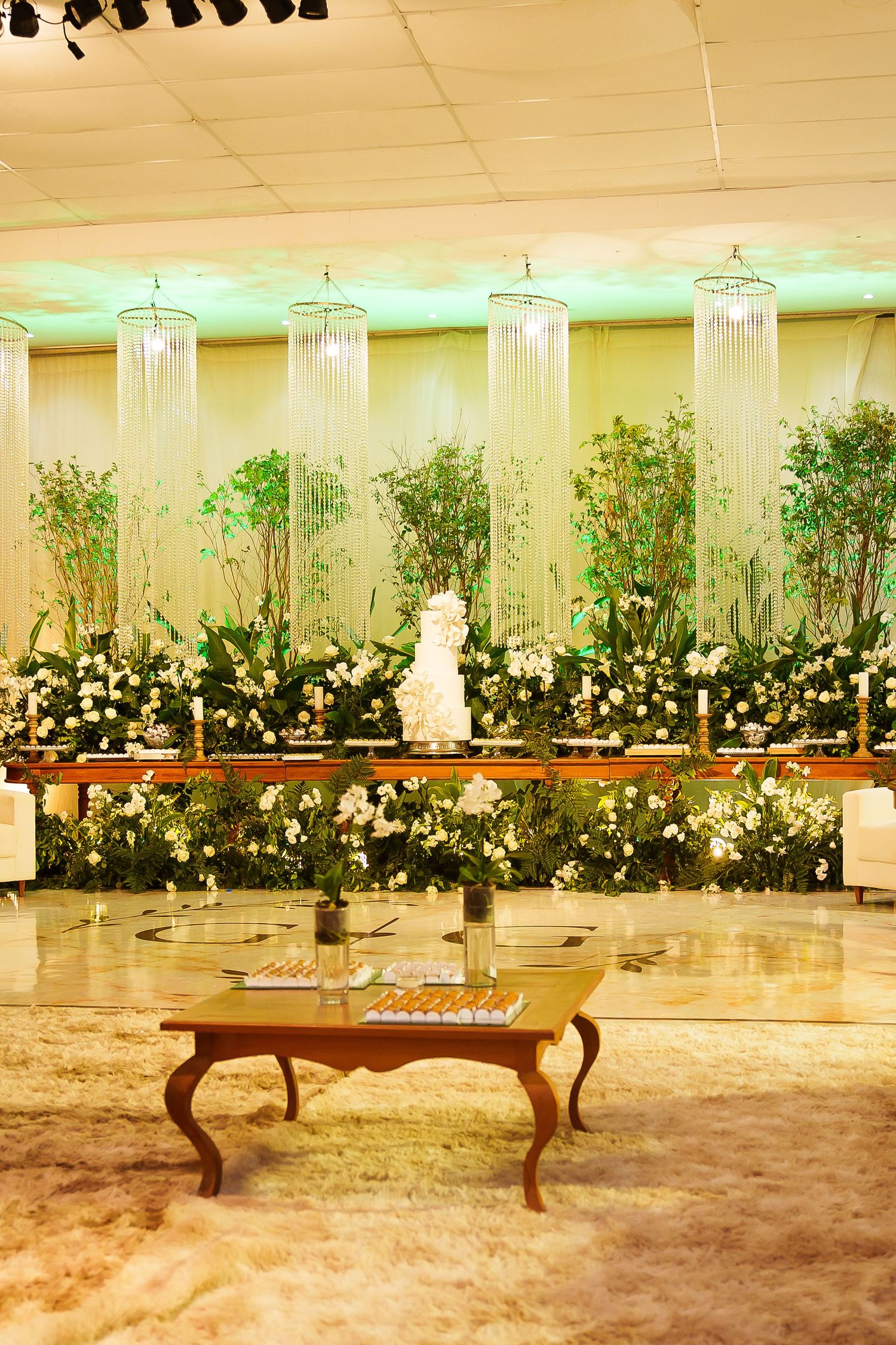 Elegant wedding reception hall with white floral arrangements, crystal chandeliers, lush greenery backdrop, and ornate wooden coffee table.