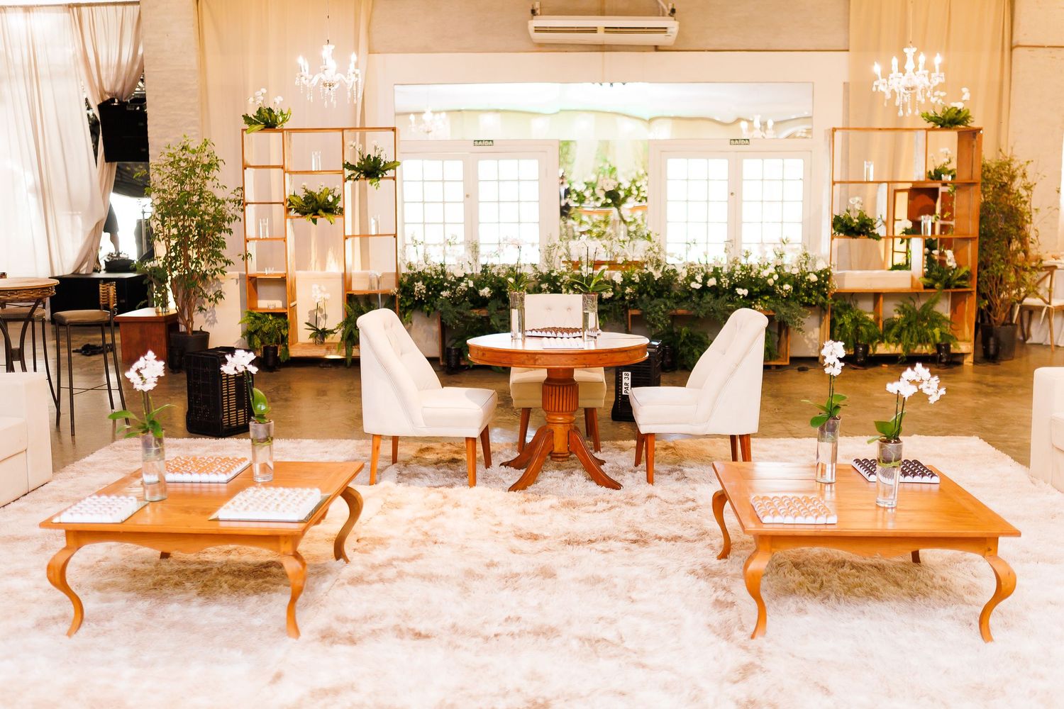 Elegant wedding reception lounge with white chairs, wooden tables, floral arrangements, and copper shelving in a bright venue.