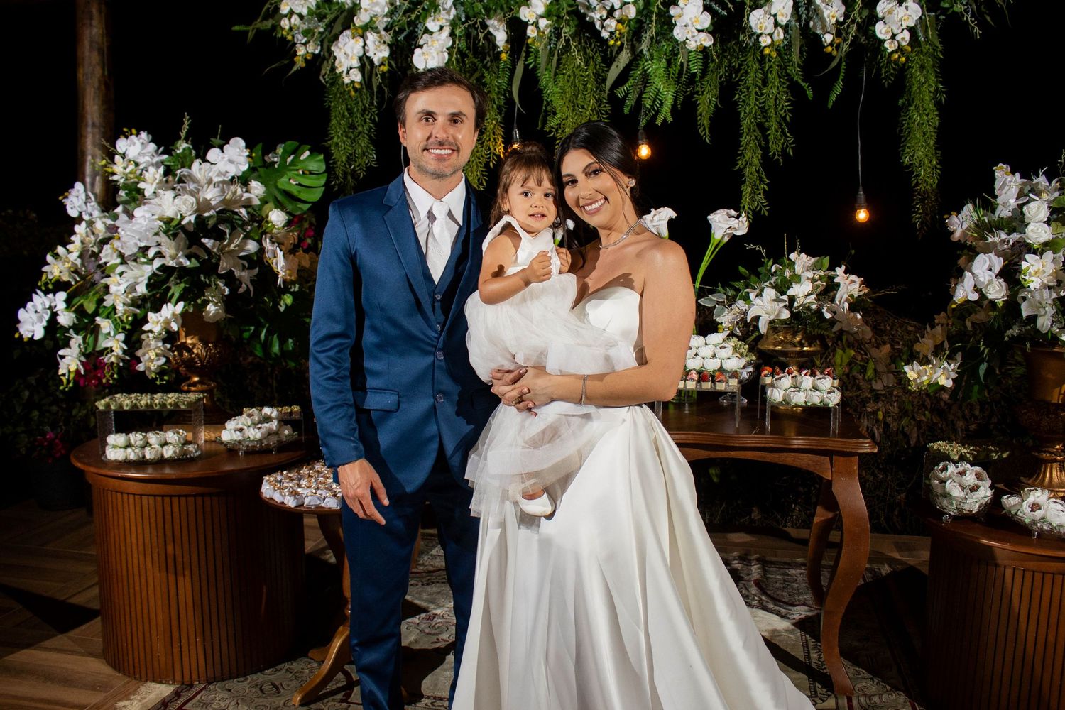 Elegant wedding couple in formal attire holding a young child, posing before a floral backdrop with white orchids.