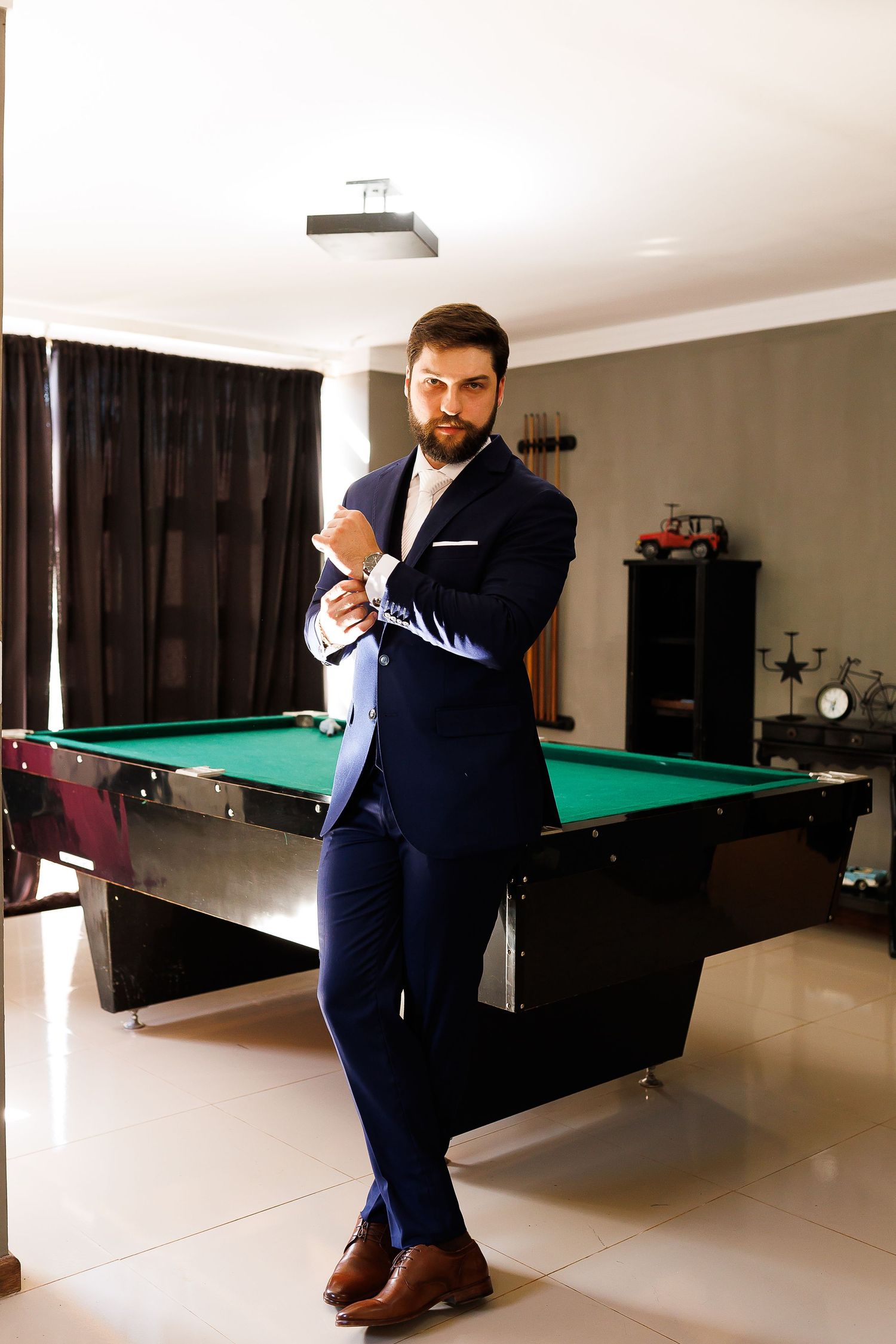 A bearded man in a navy blue suit stands confidently beside a green billiard table in an elegant room.