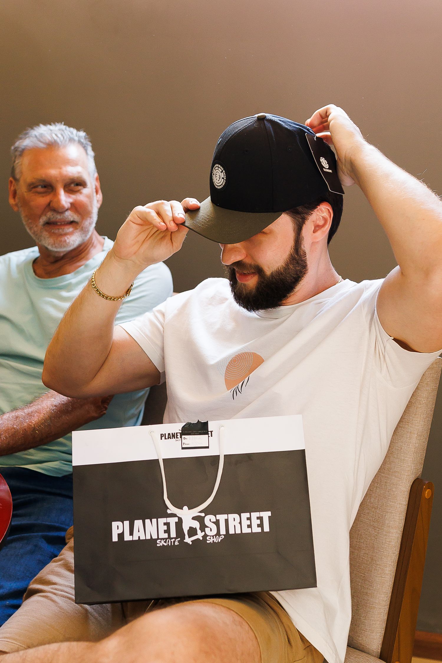 Man trying on black cap from Planet Street Skate Shop bag, with older man smiling in background.