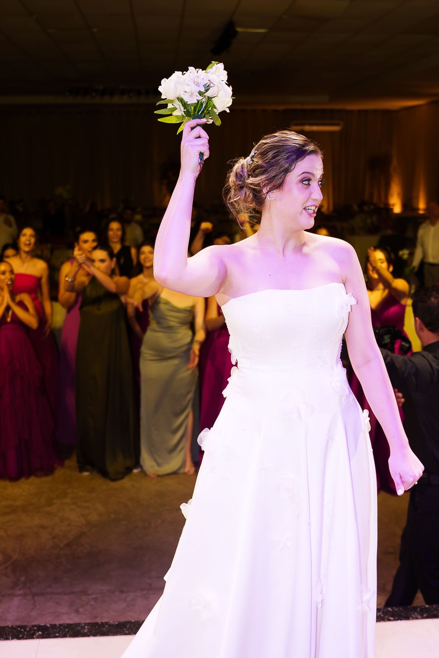 Bride in white strapless gown raises bouquet of white flowers during wedding reception bouquet toss.