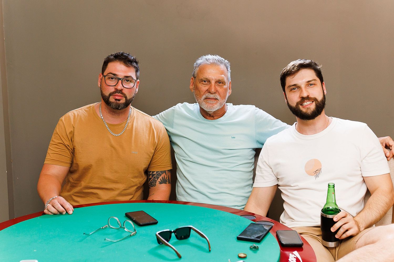 Three smiling men pose together at a green poker table with drinks and sunglasses nearby.