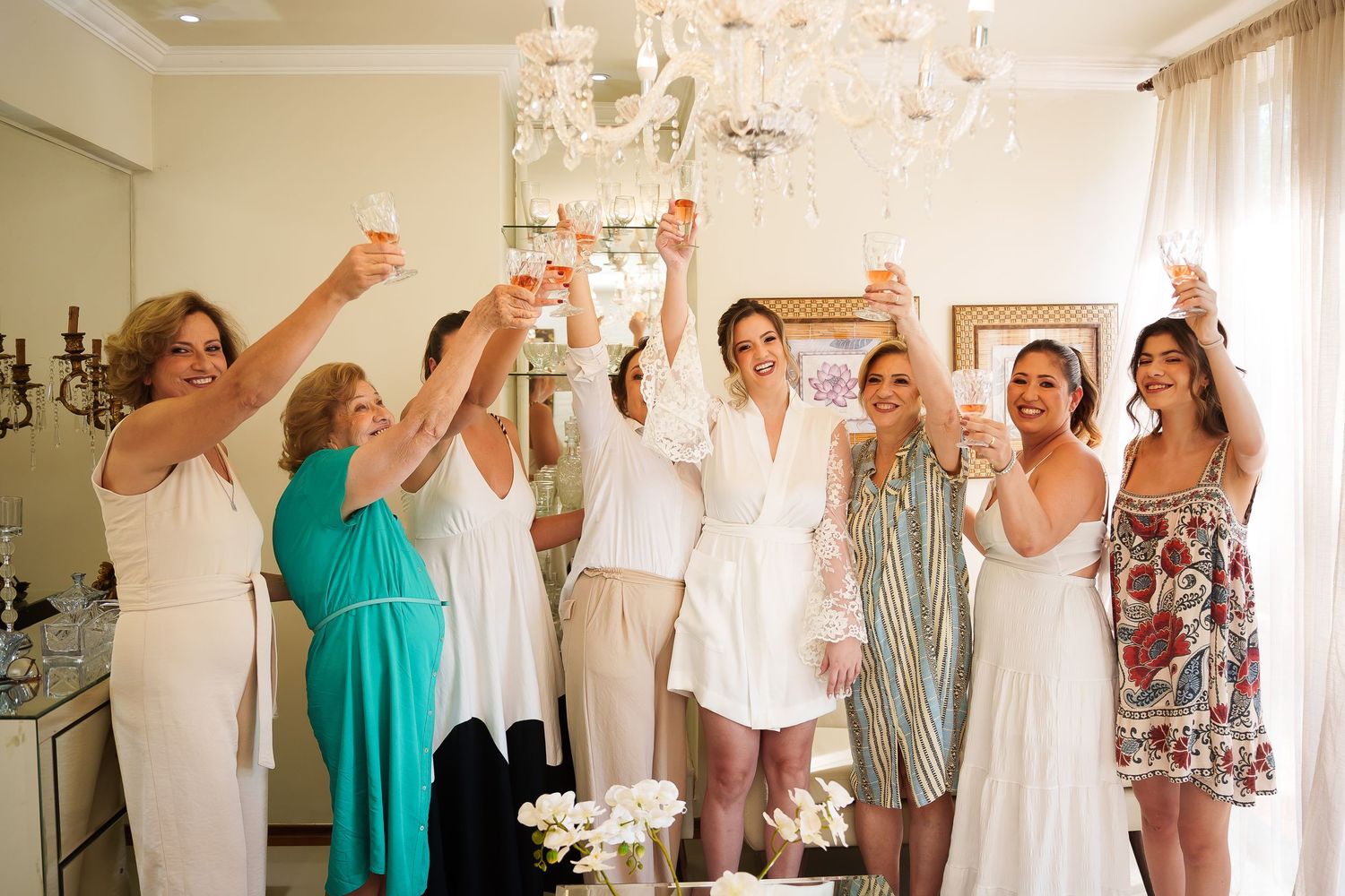 Group of women in elegant attire toasting with champagne glasses under a chandelier at a bridal celebration.