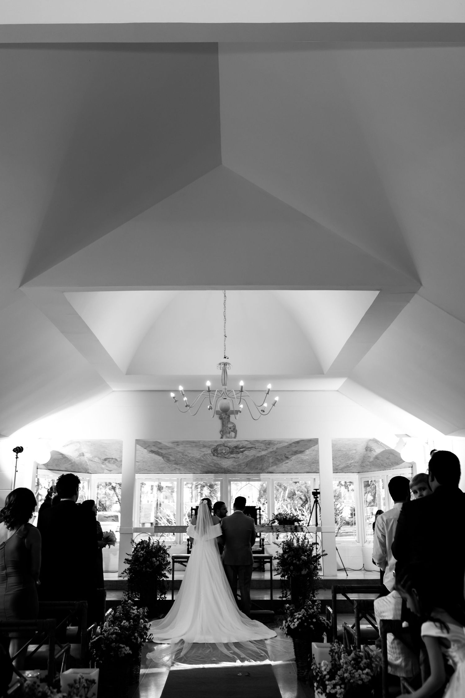 Black and white photo of a bride walking down the aisle in a chapel with guests standing and a chandelier above.