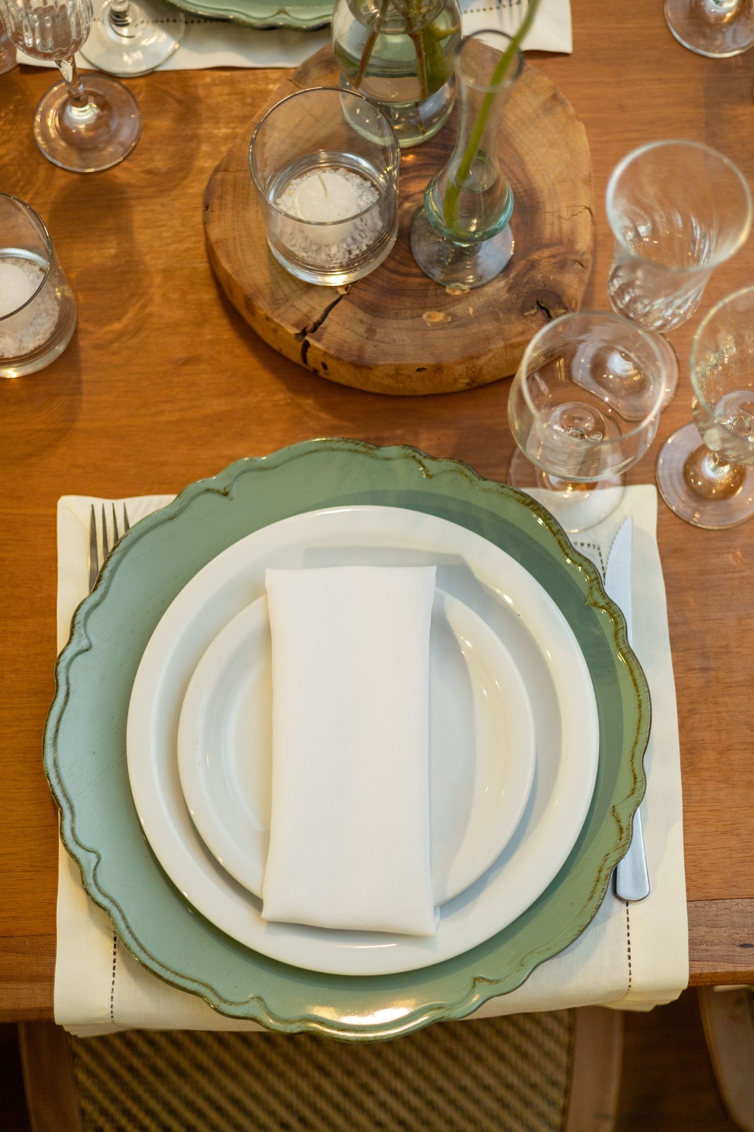 Elegant table setting with green charger plate, white dinner plate, folded napkin, crystal glasses, and wooden centerpiece.