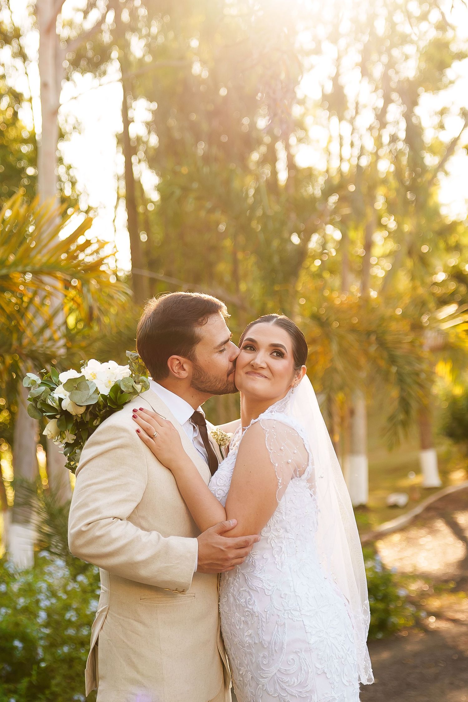 Groom in beige suit kisses bride in white lace dress outdoors, surrounded by lush green trees in golden sunlight.