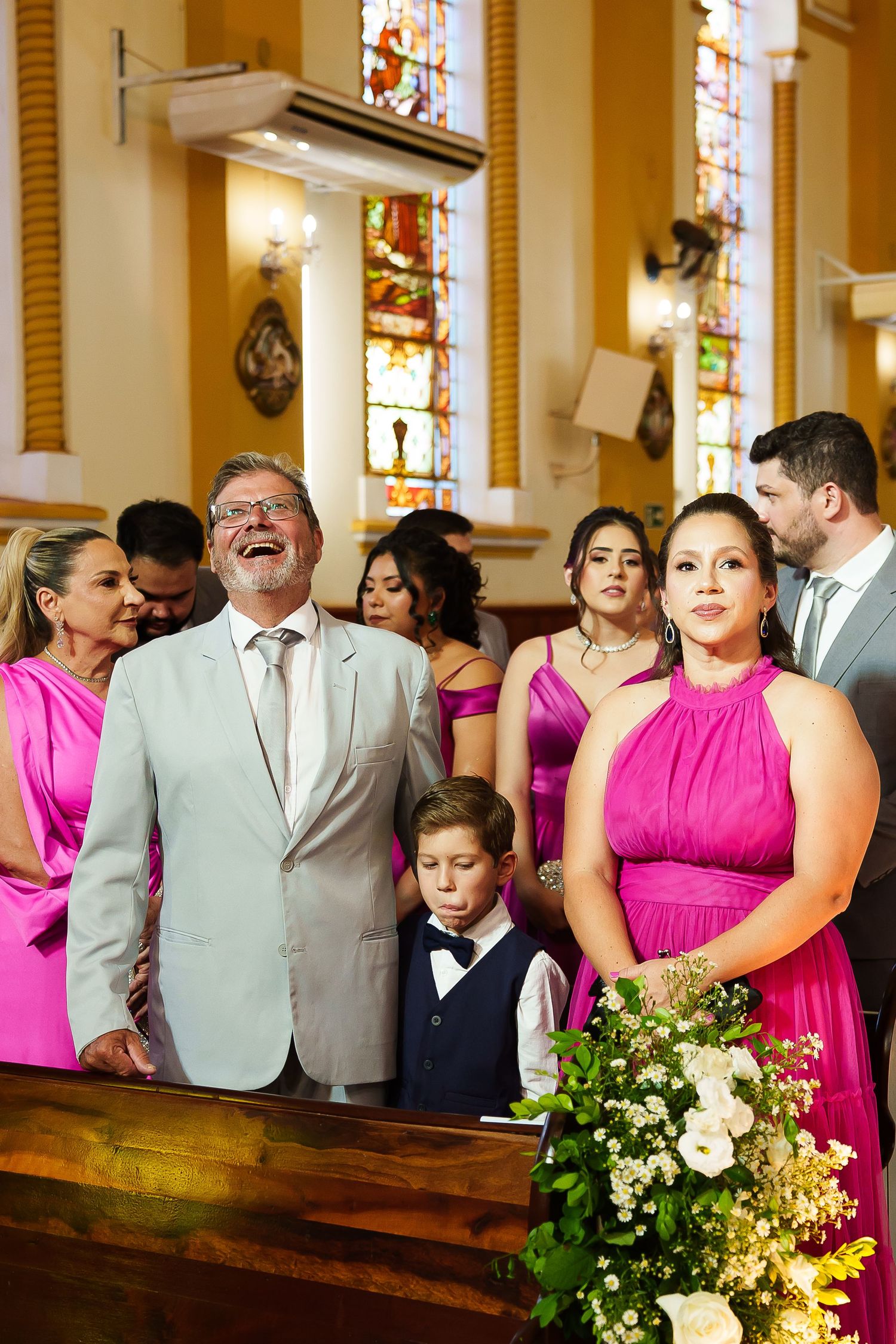 Wedding party in pink dresses and suits laughing joyfully inside an ornate church with stained glass windows.