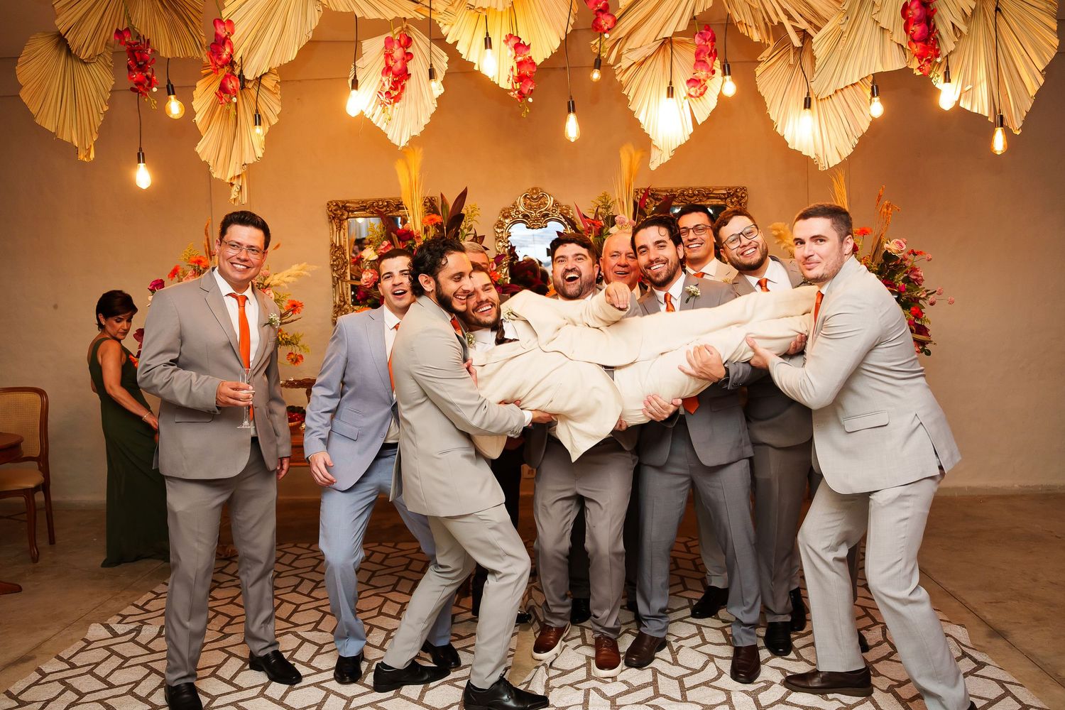 Groomsmen in gray suits lift the groom during a fun wedding celebration under tropical floral decorations.