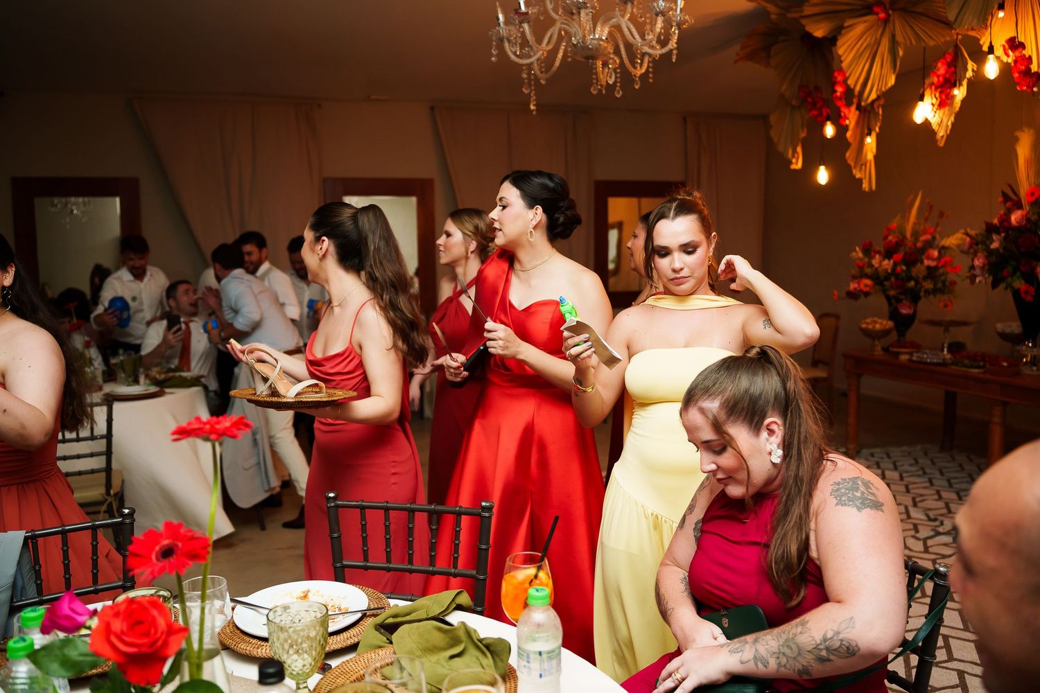 Bridesmaids in red and yellow dresses mingle at a wedding reception with floral decorations and elegant chandelier lighting.