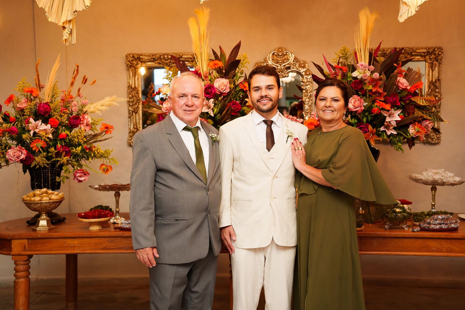 A young man in a white suit poses with an older couple in elegant attire at a floral-decorated wedding reception.