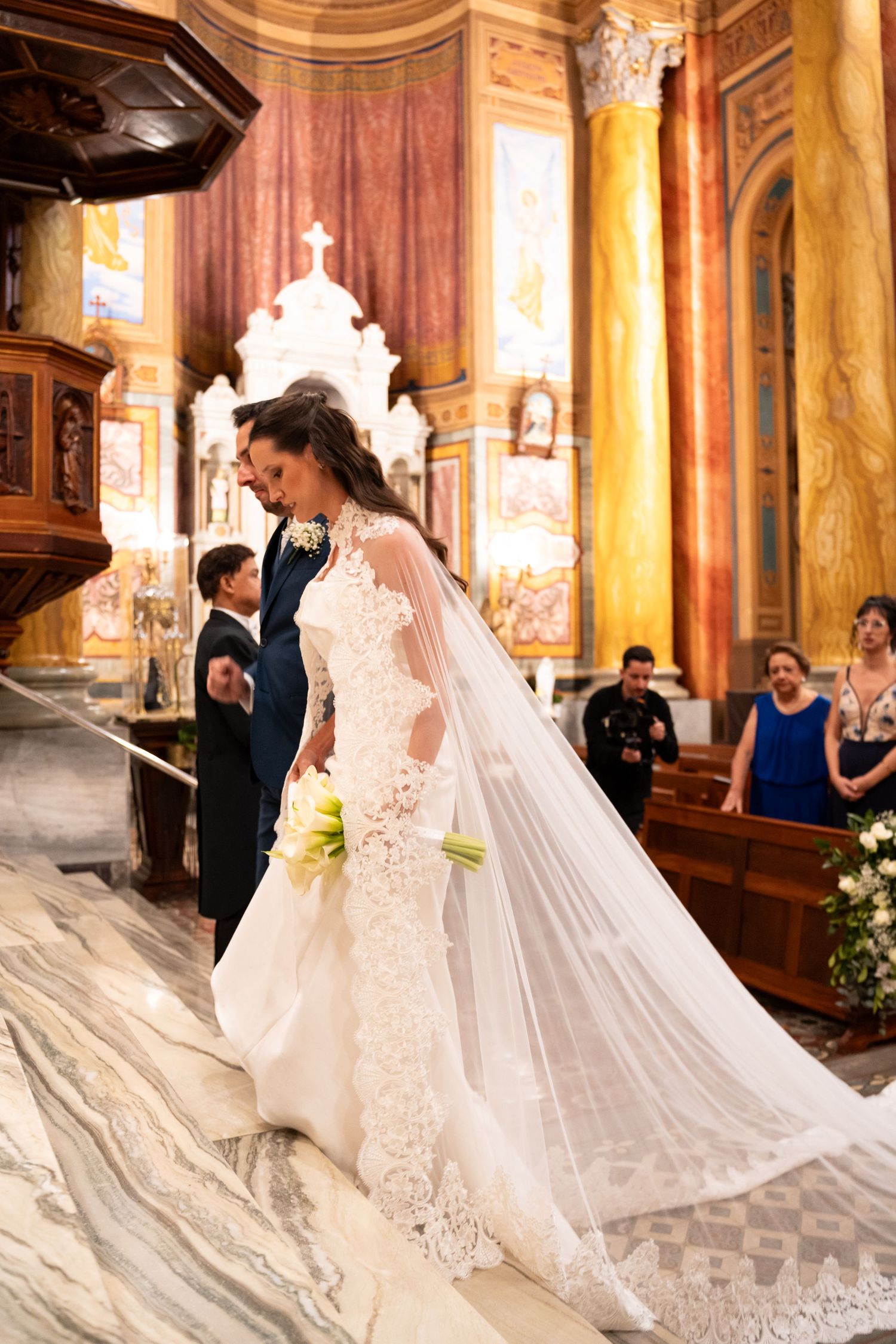 Bride in lace gown with cathedral veil walks church aisle holding bouquet, groom and guests in ornate Italian church.