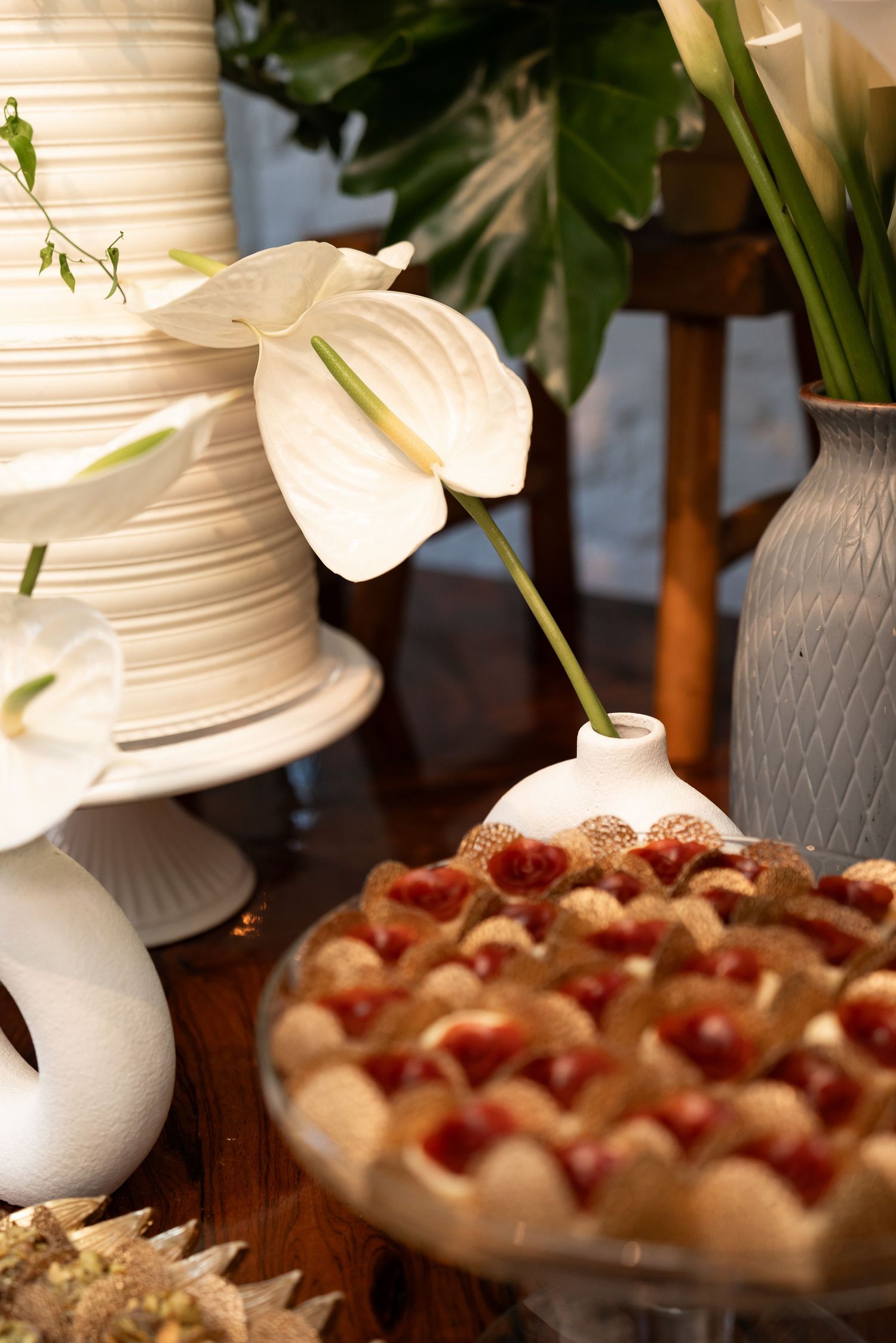 Elegant dessert table with white anthurium flowers, tiered wedding cake, and cherry tart pastries on wooden surface.