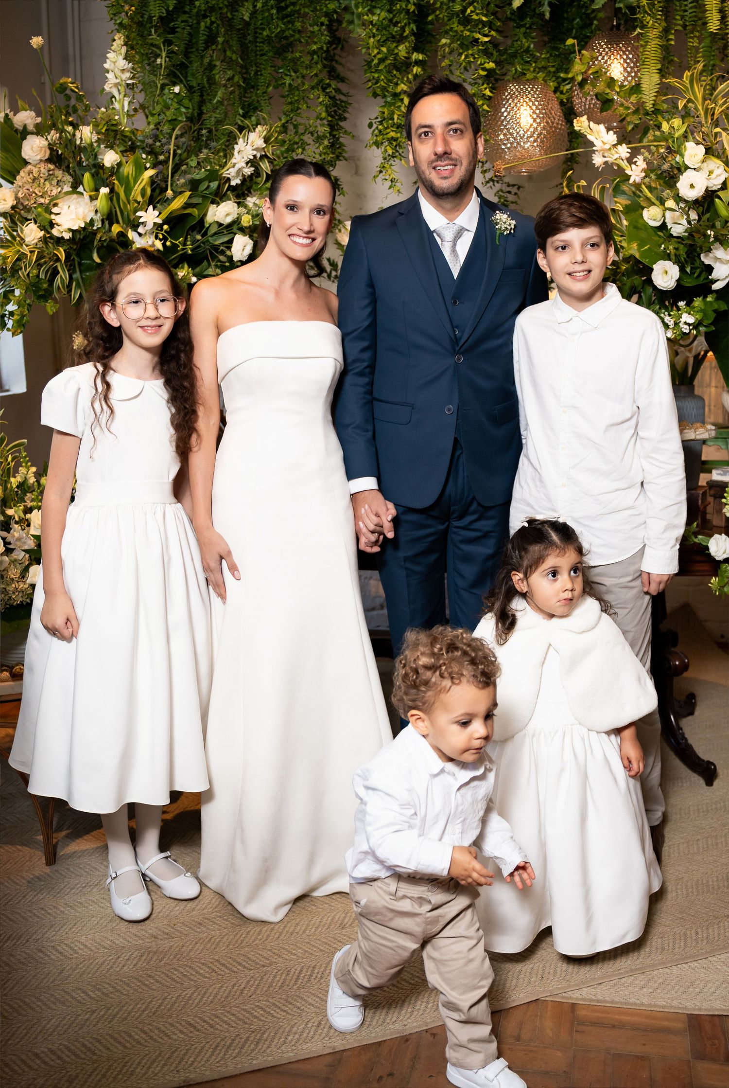 Bride and groom pose with four children in white attire at an elegant wedding ceremony with floral decor.