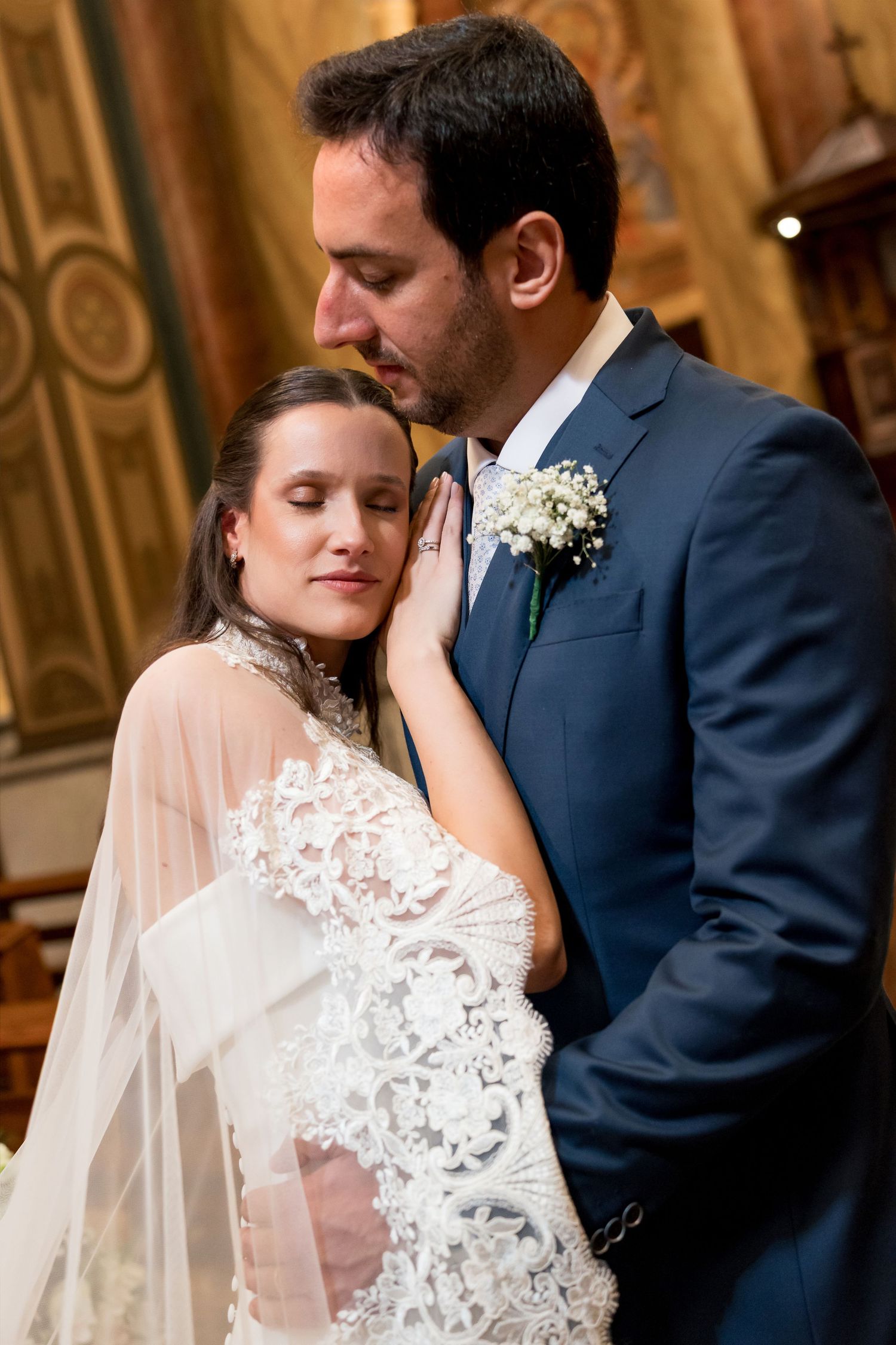 A groom in a navy suit kisses his bride's forehead as she leans against him in a lace wedding gown inside a church.