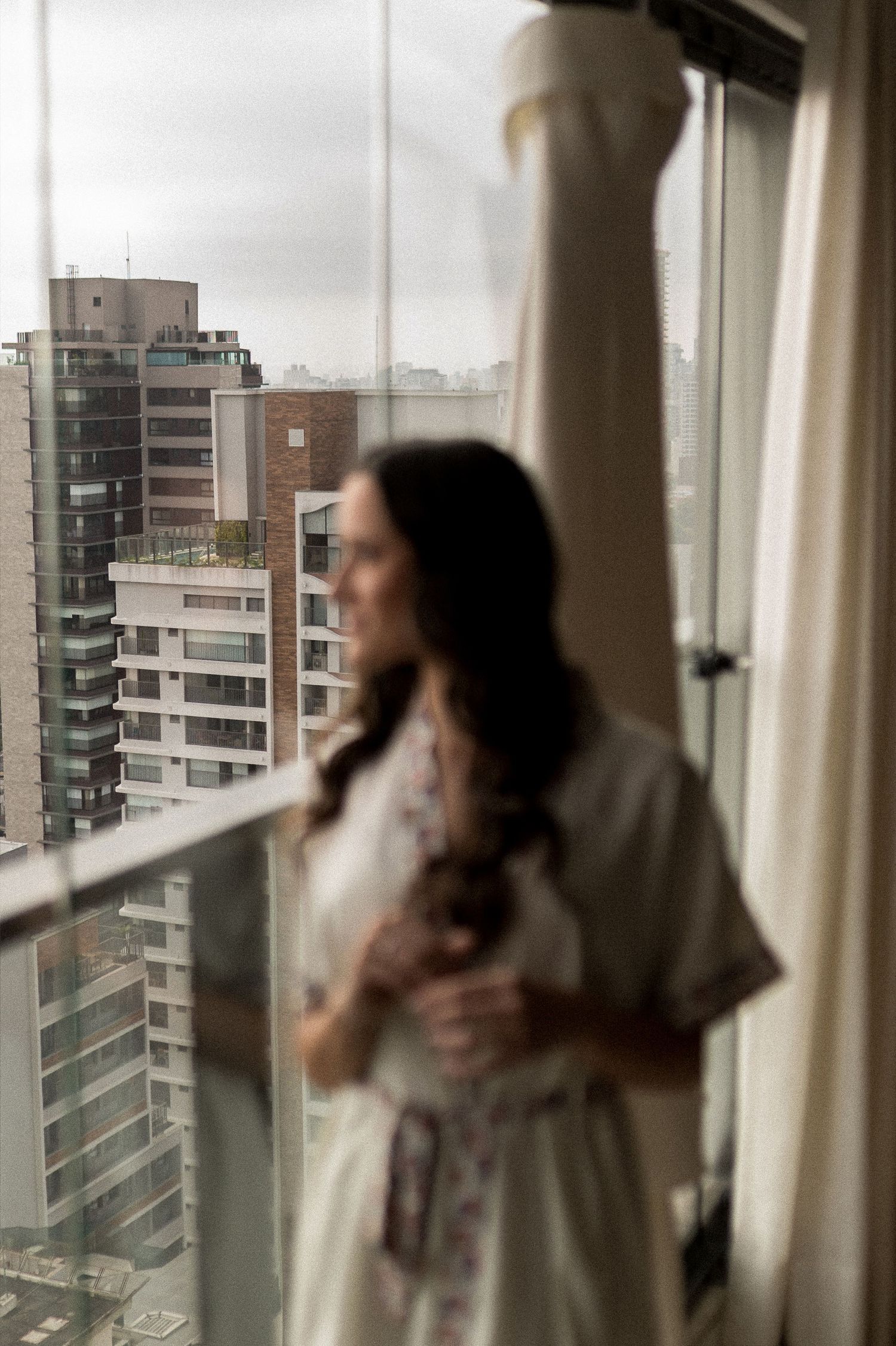 Bride in white robe gazes out hotel window at city skyline, wedding dress hanging nearby.