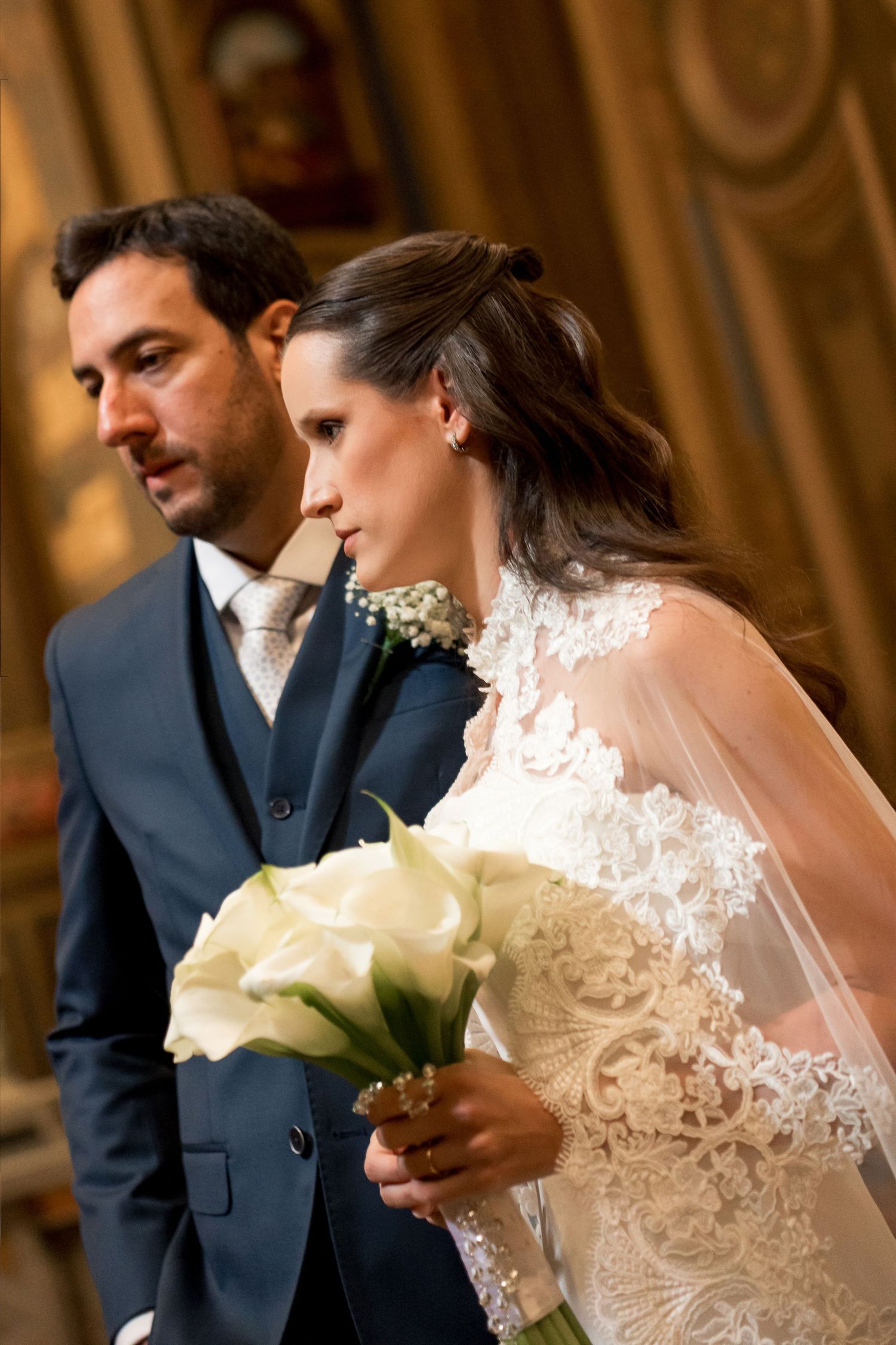 Bride in lace gown holding white calla lily bouquet stands beside groom in navy suit during wedding ceremony.