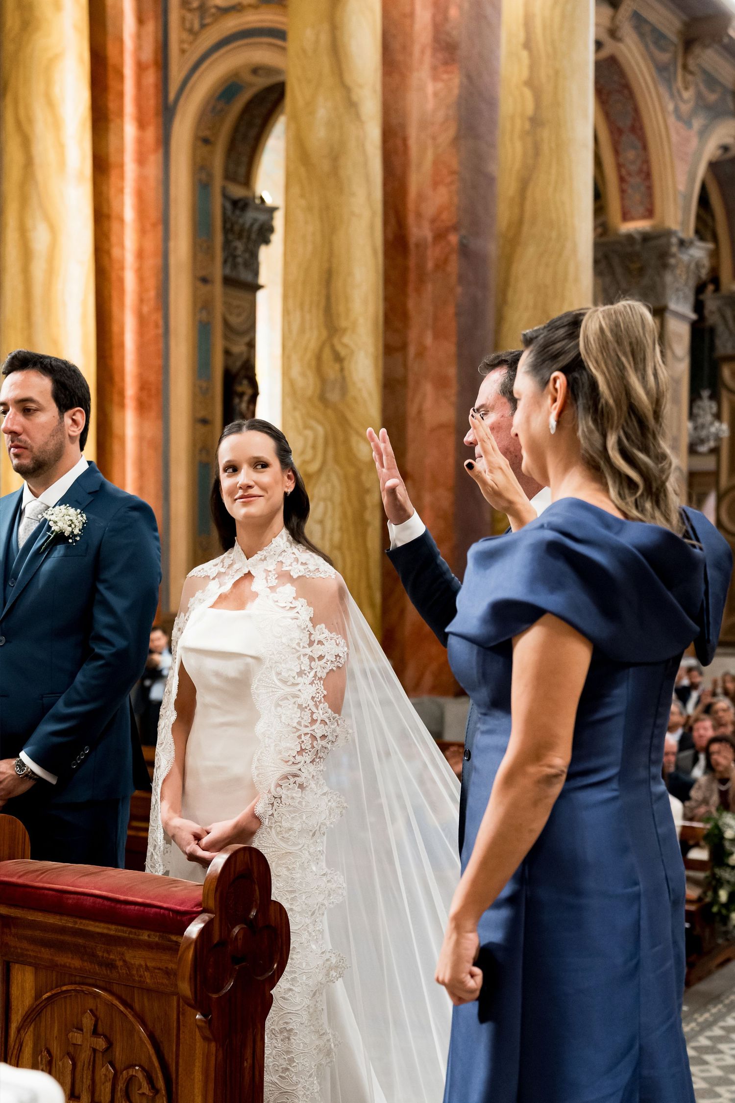 Bride in lace wedding gown stands at church altar during ceremony while guests in blue attire gesture nearby.