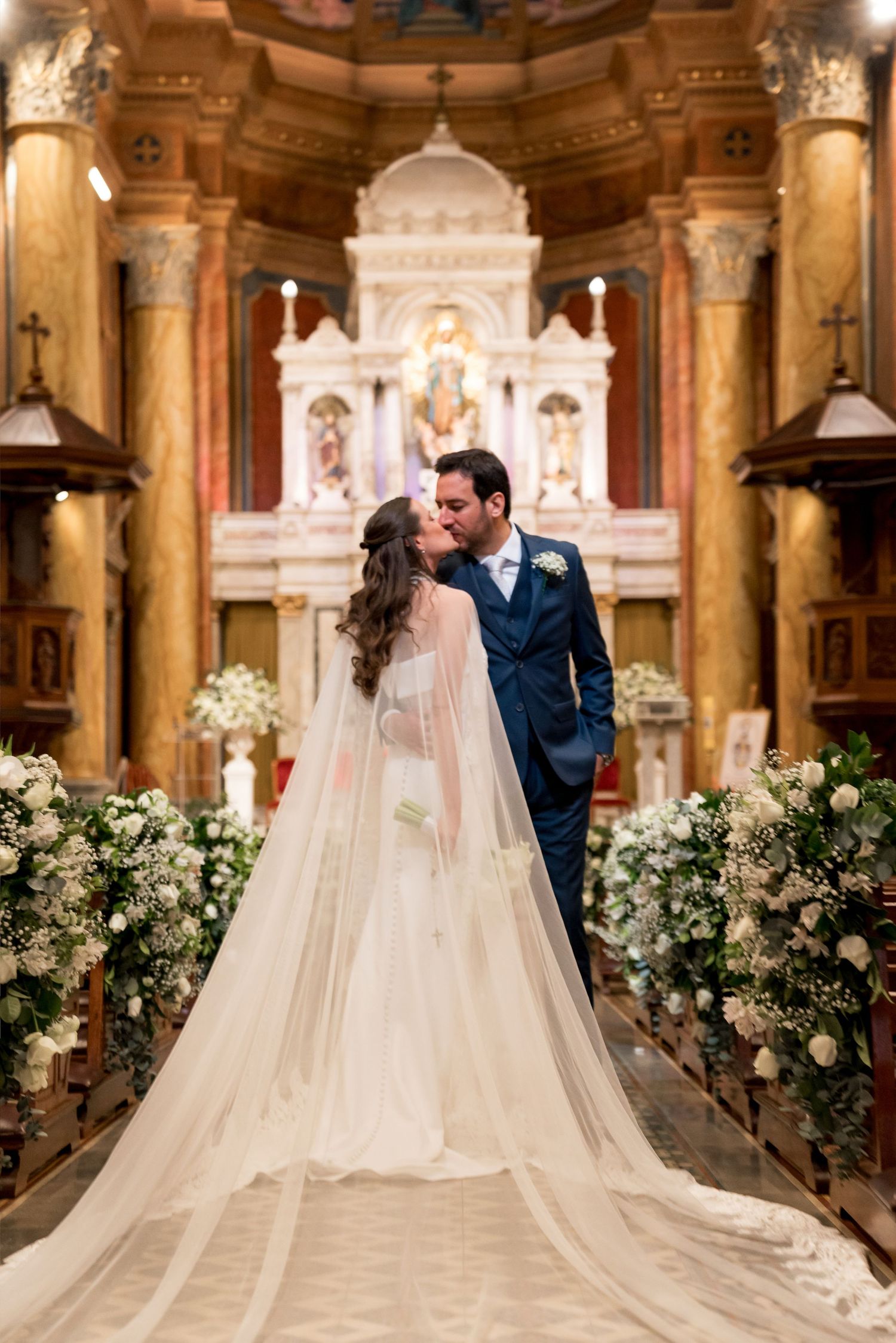 Bride and groom share a kiss at the altar of an ornate church, surrounded by white floral arrangements.