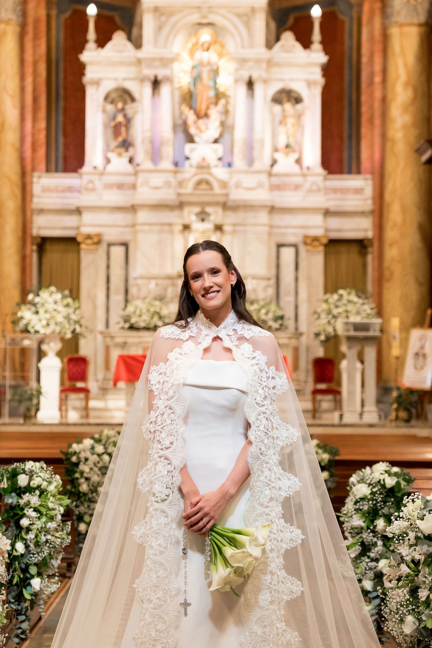 Bride in white gown and lace cape veil holding calla lily bouquet stands before ornate church altar.