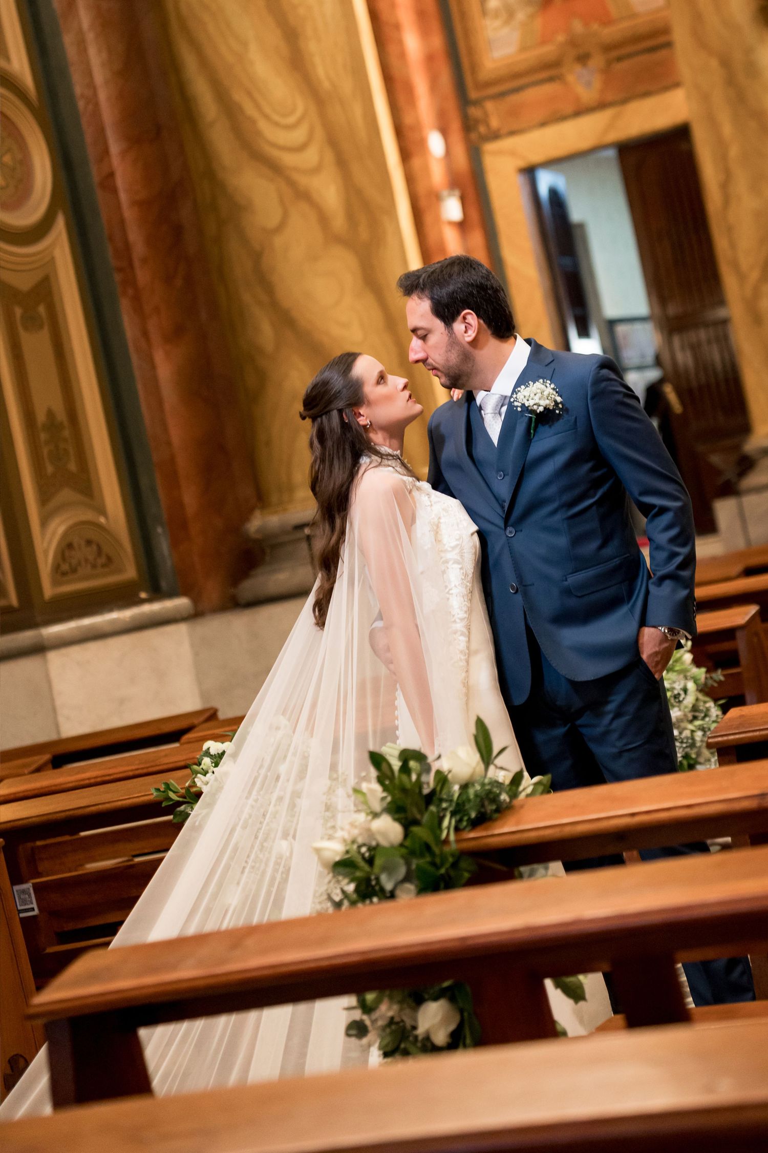 Bride and groom share a romantic kiss in a church aisle, surrounded by wooden pews and floral decorations.