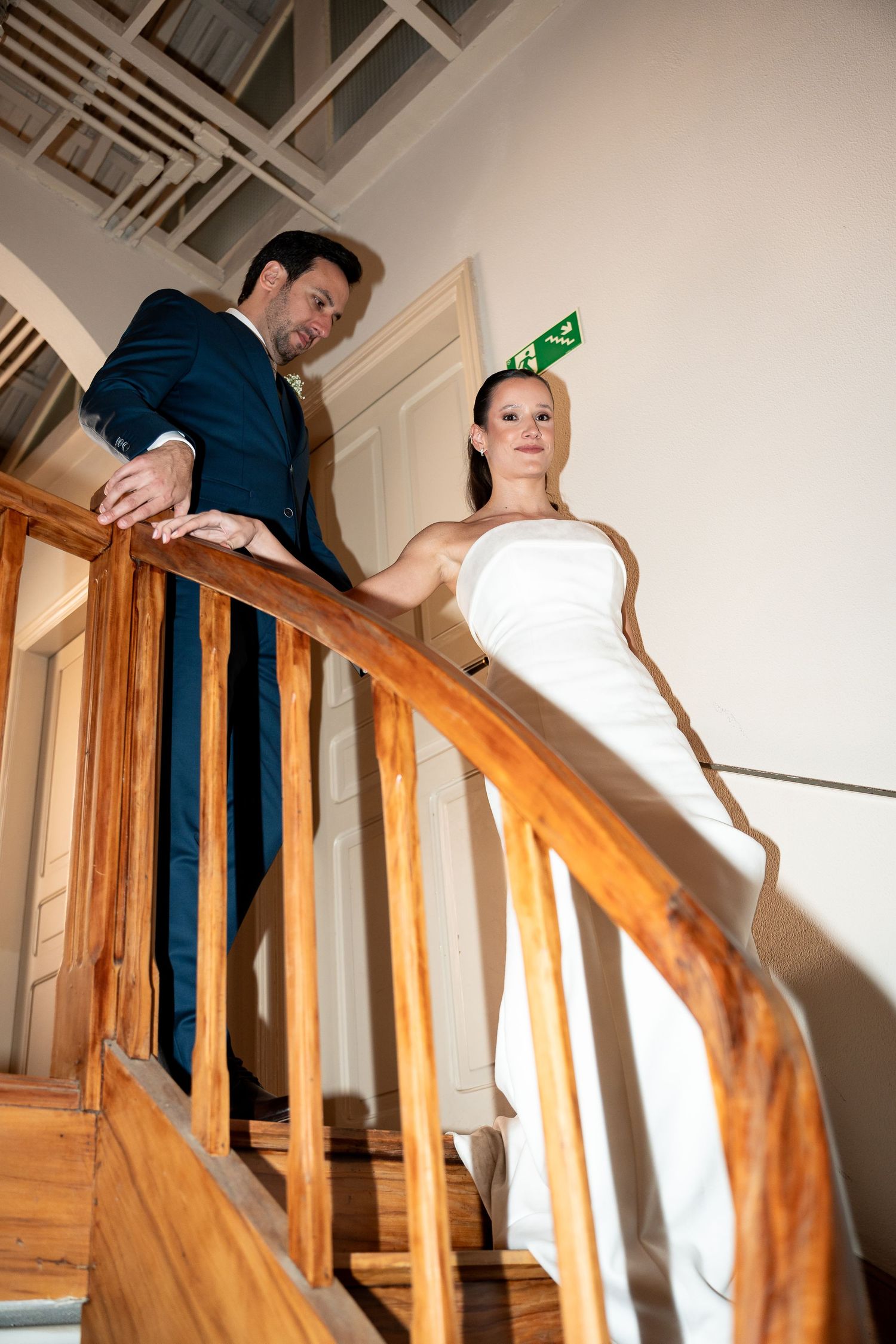 Bride in white gown and groom in navy suit pose on wooden staircase in elegant indoor wedding venue.