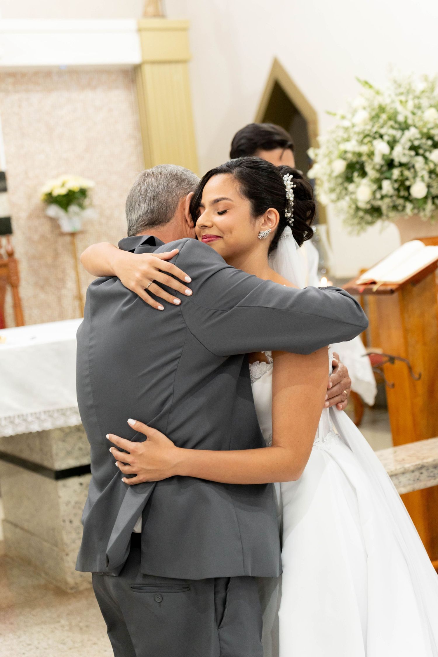 Bride in white gown hugging man in gray suit at church altar during wedding ceremony.
