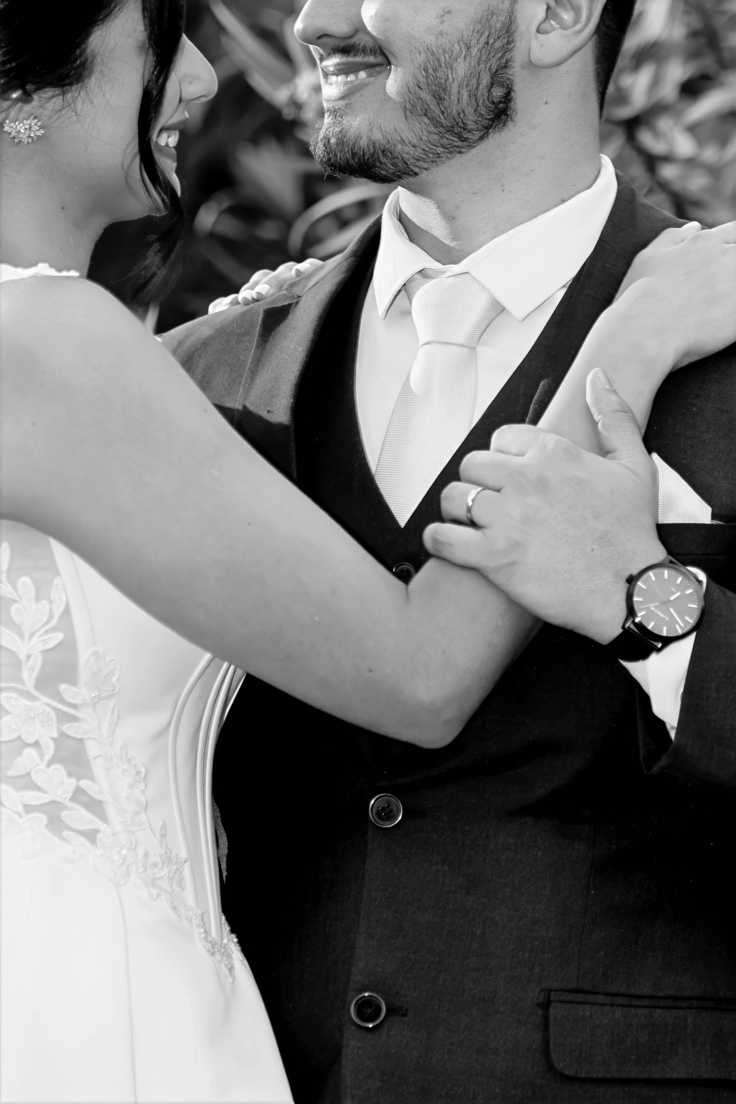 Black and white wedding photo of smiling groom in suit embracing bride in lace dress, both wearing rings.