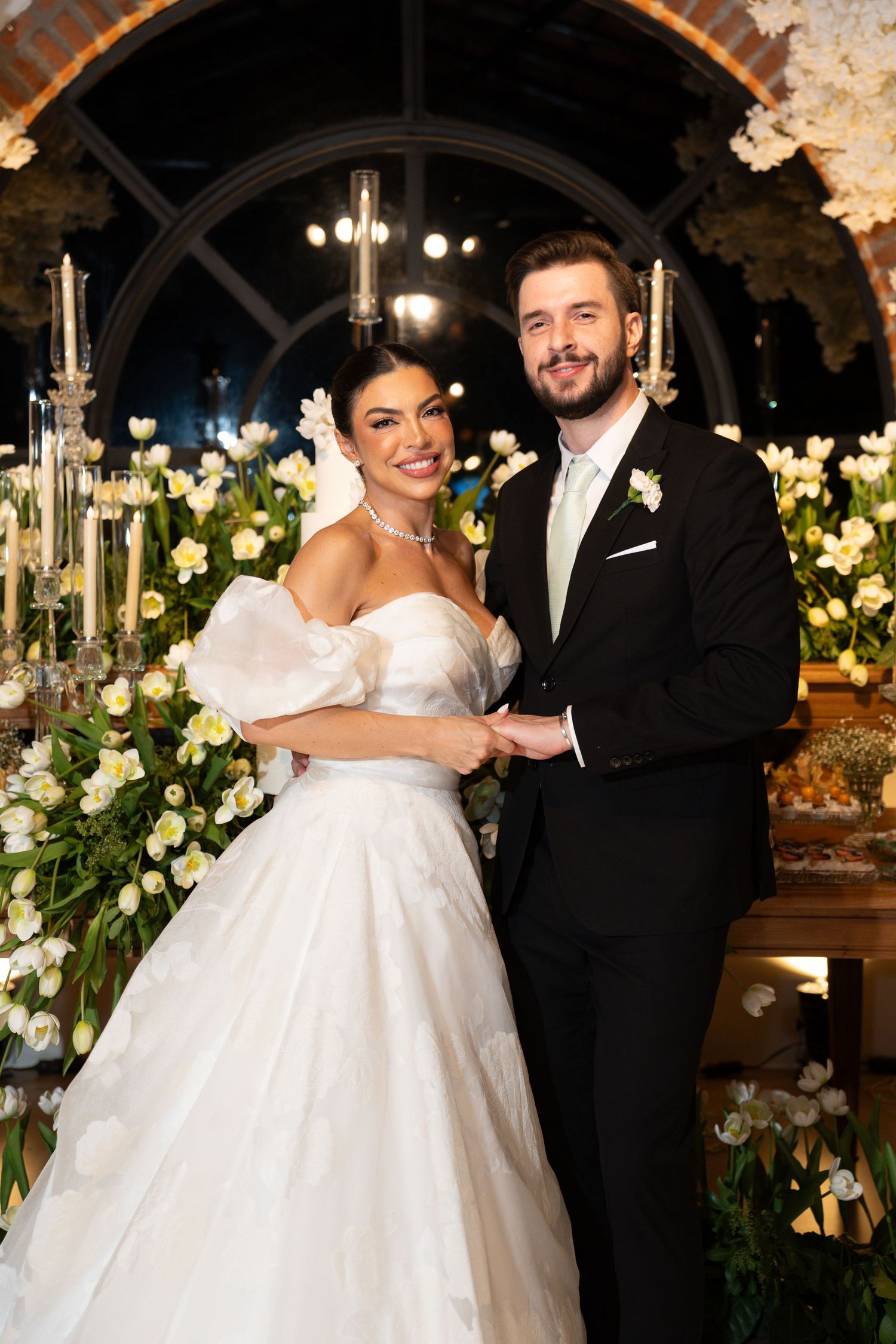 Bride in white ballgown and groom in black tuxedo holding hands, smiling before floral wedding altar.