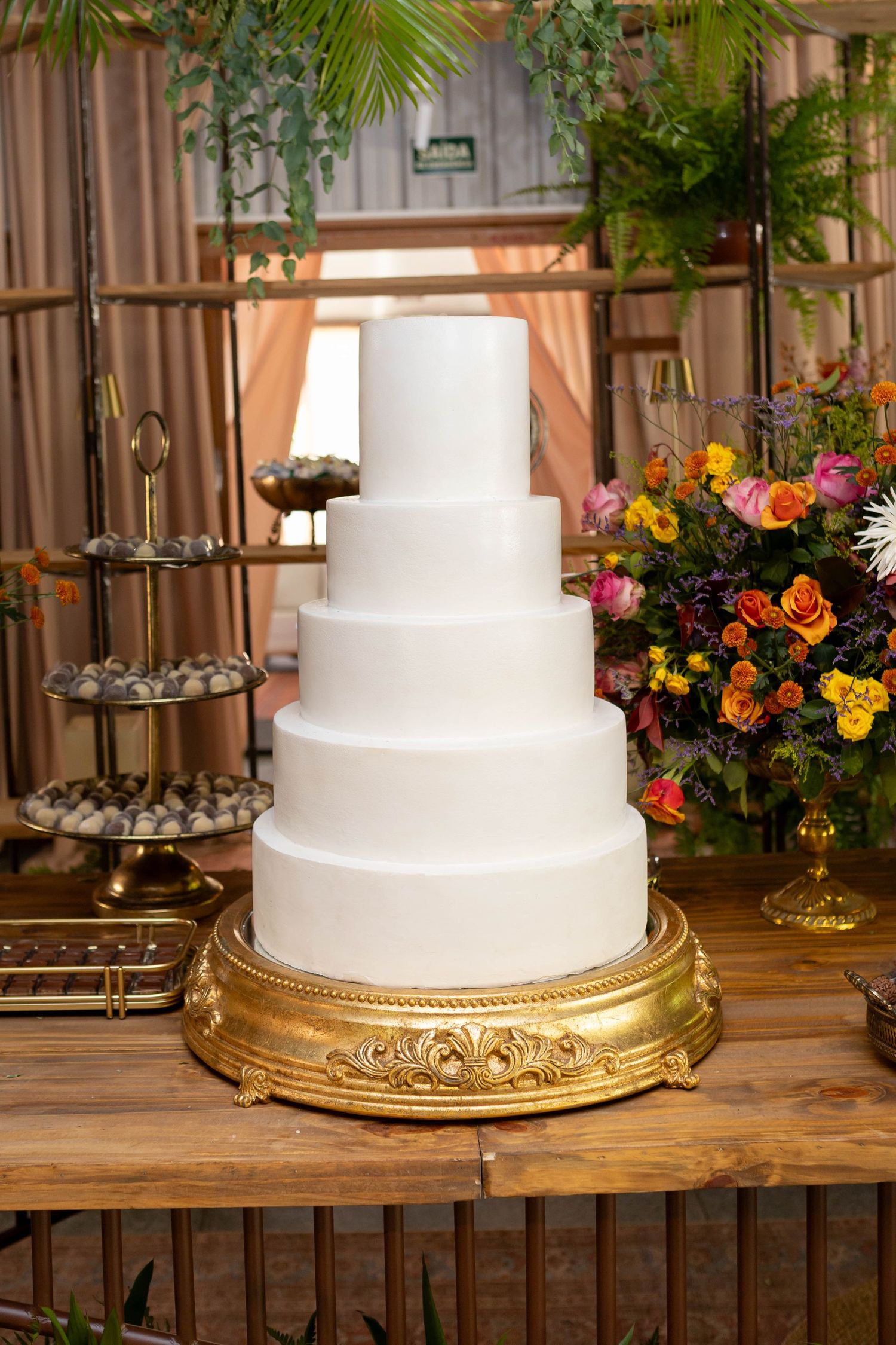 Elegant five-tier white wedding cake on a gold stand, surrounded by colorful floral arrangements on a wooden table.