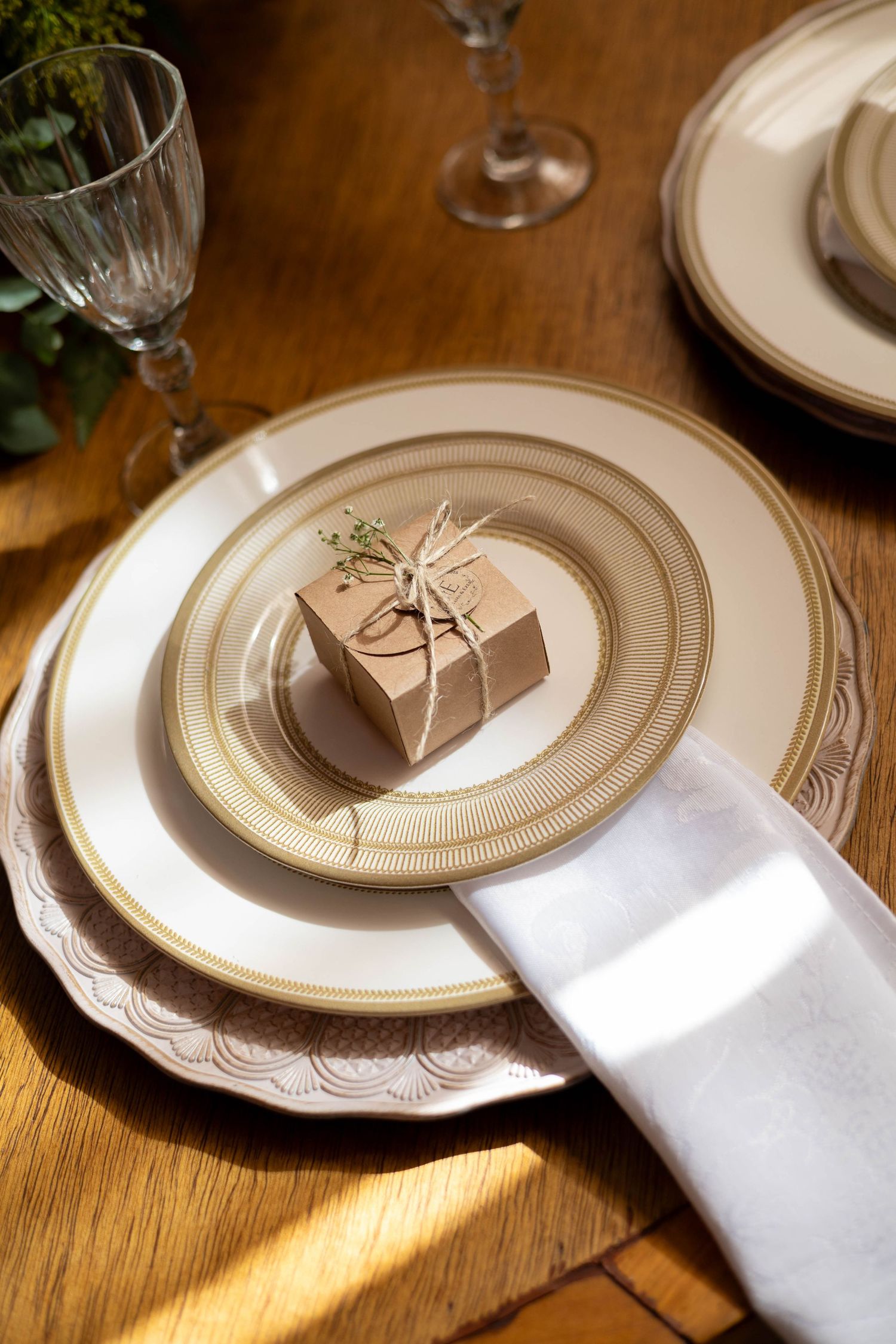 Elegant table setting with gold-rimmed plates, white napkin, kraft paper gift box tied with twine, and crystal glassware on wood table.