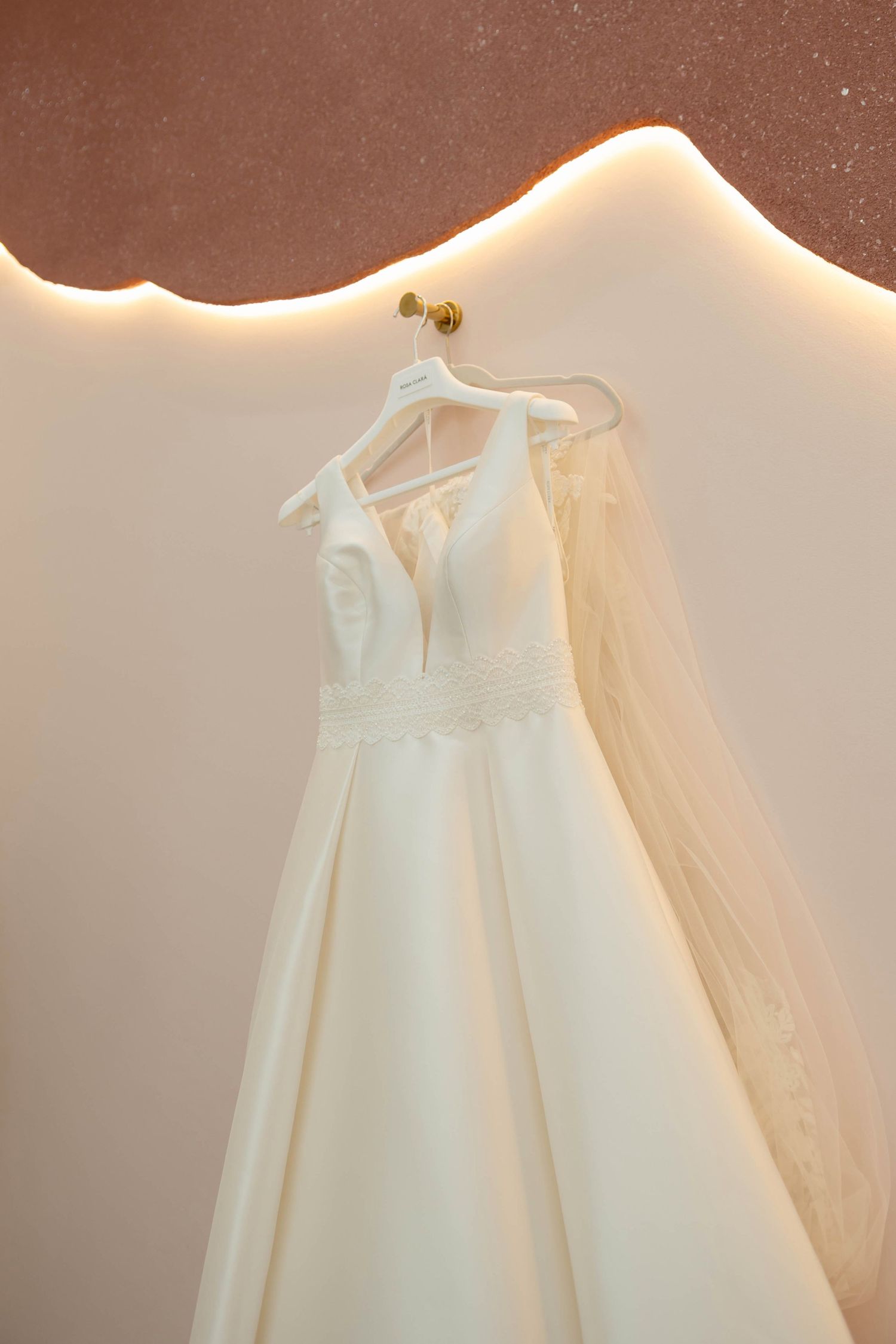 Elegant white A-line wedding dress with V-neckline and lace waist detail hanging on gold hook against warm beige wall.