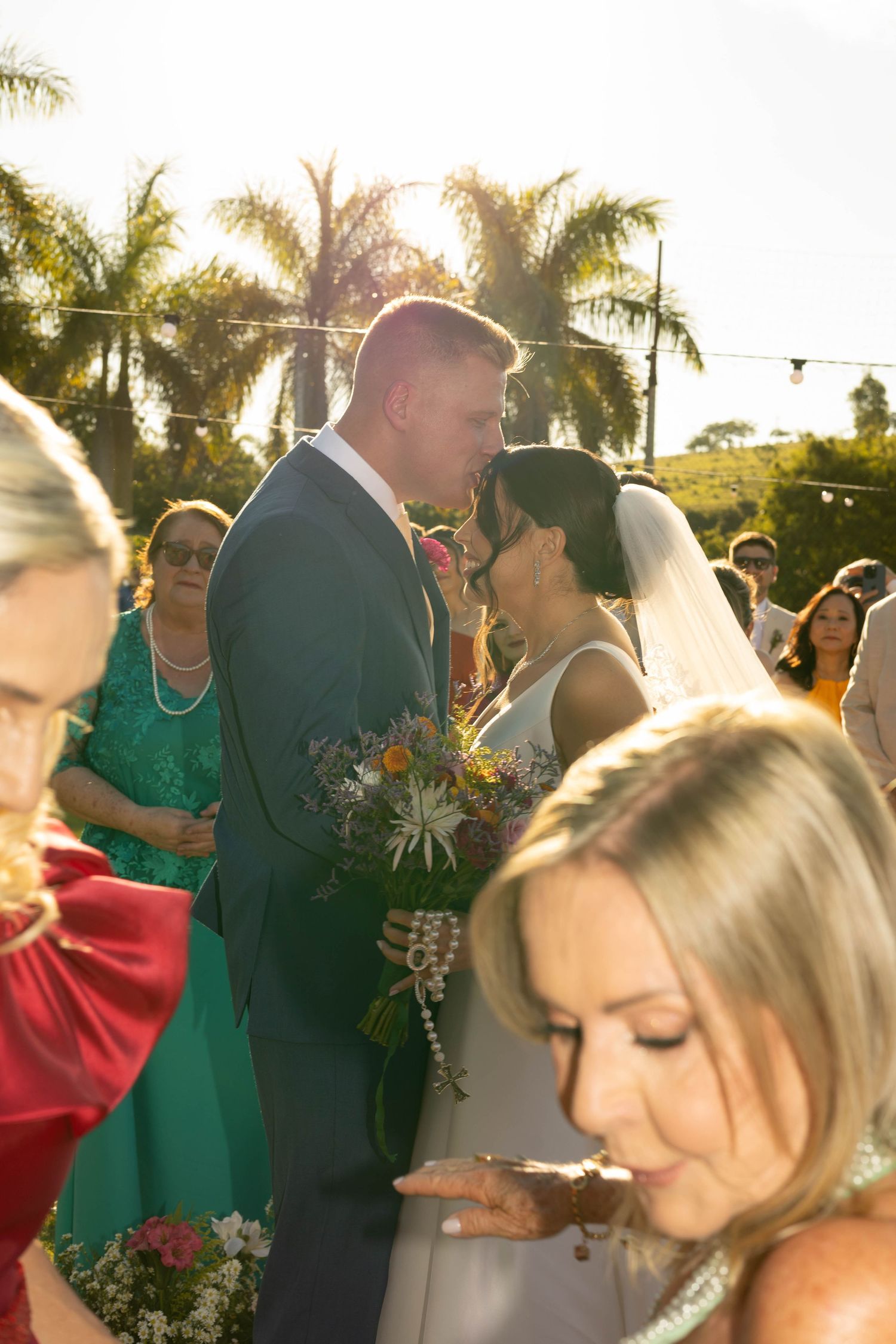 Bride and groom share a kiss at outdoor wedding ceremony with guests and palm trees in golden sunlight.