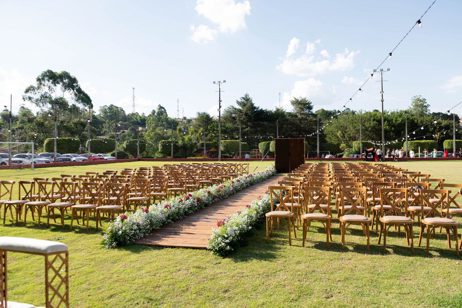 Outdoor wedding ceremony setup with wooden chairs lining a floral aisle on green lawn with string lights overhead.