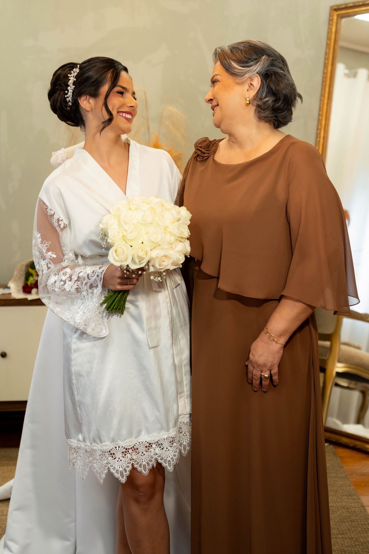 Bride in white lace robe holding bouquet of white roses smiles with older woman in brown dress in elegant room.