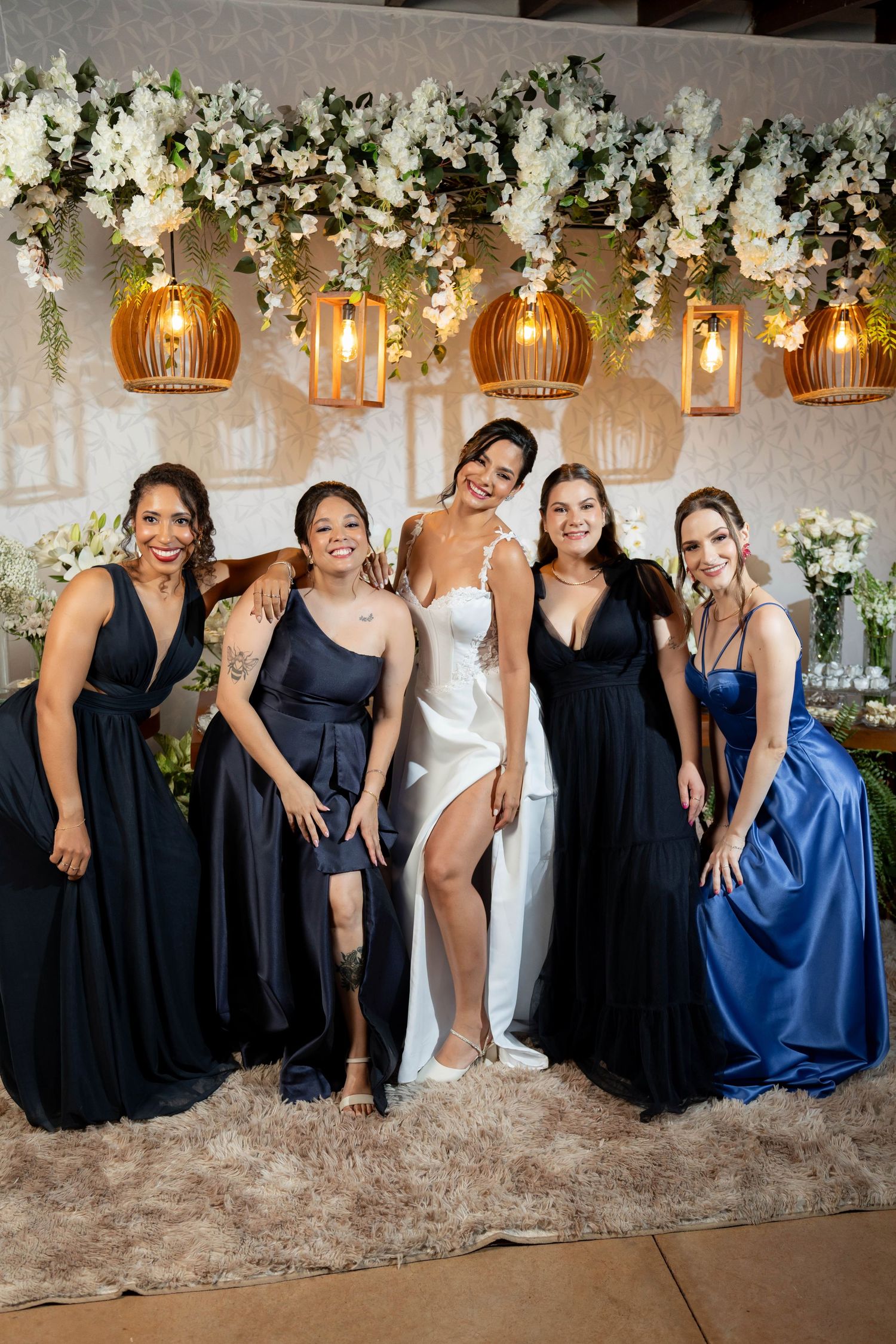 Bride in white gown poses with four bridesmaids in navy and blue dresses under floral ceiling decor.