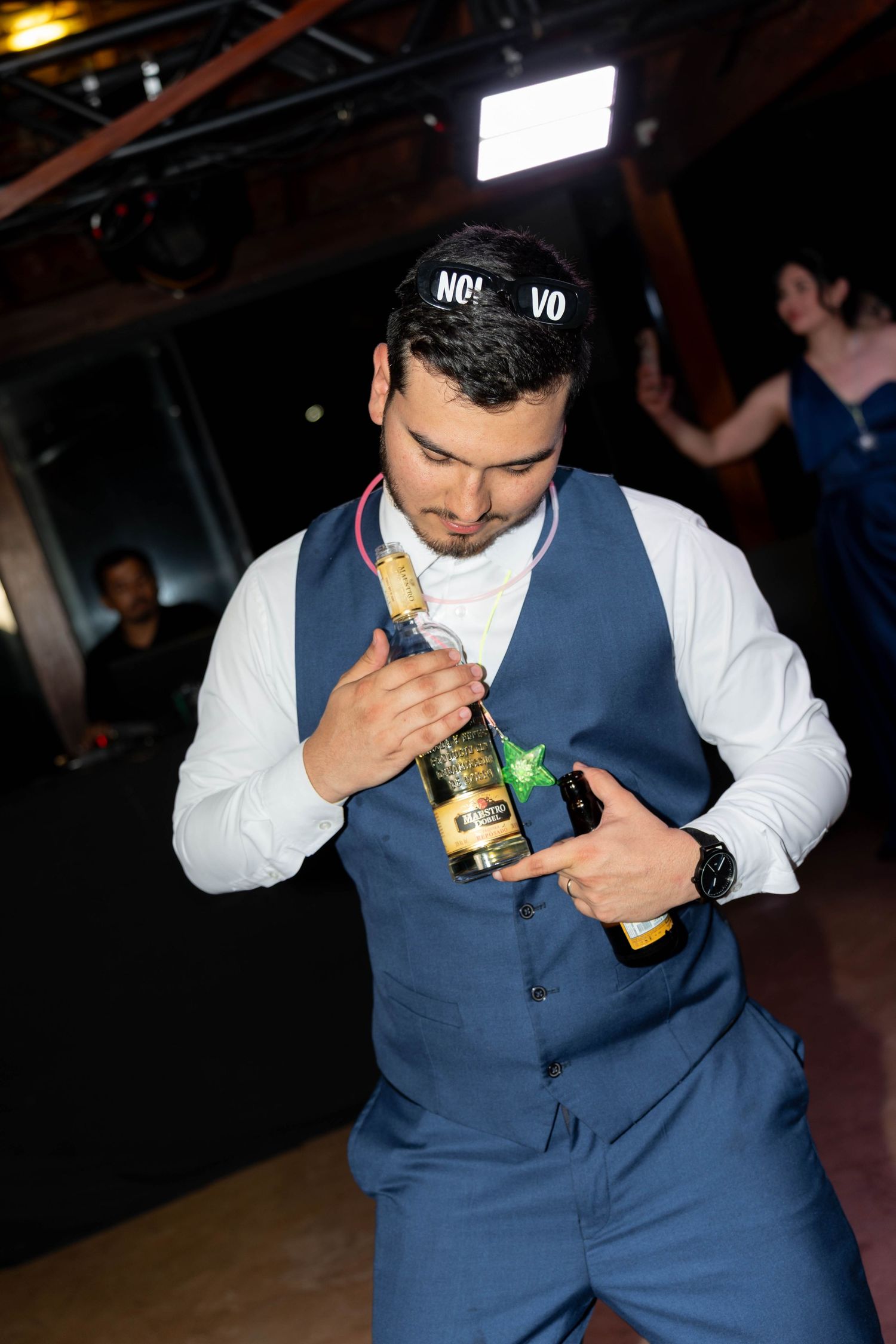 Man in blue vest holding Corona beer bottles while wearing a New Year's party hat at a celebration event.
