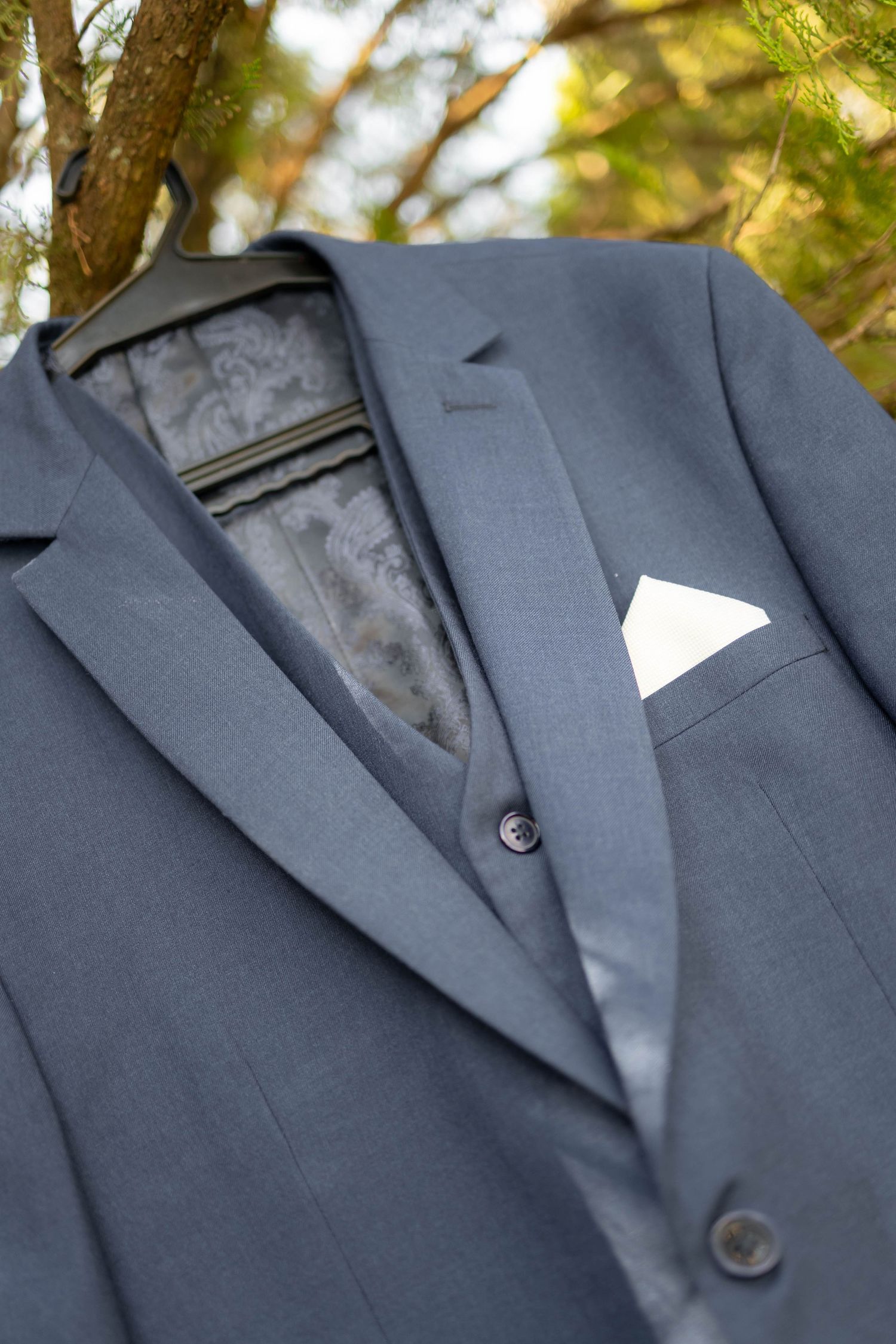 Gray men's suit jacket with white pocket square on hanger outdoors.